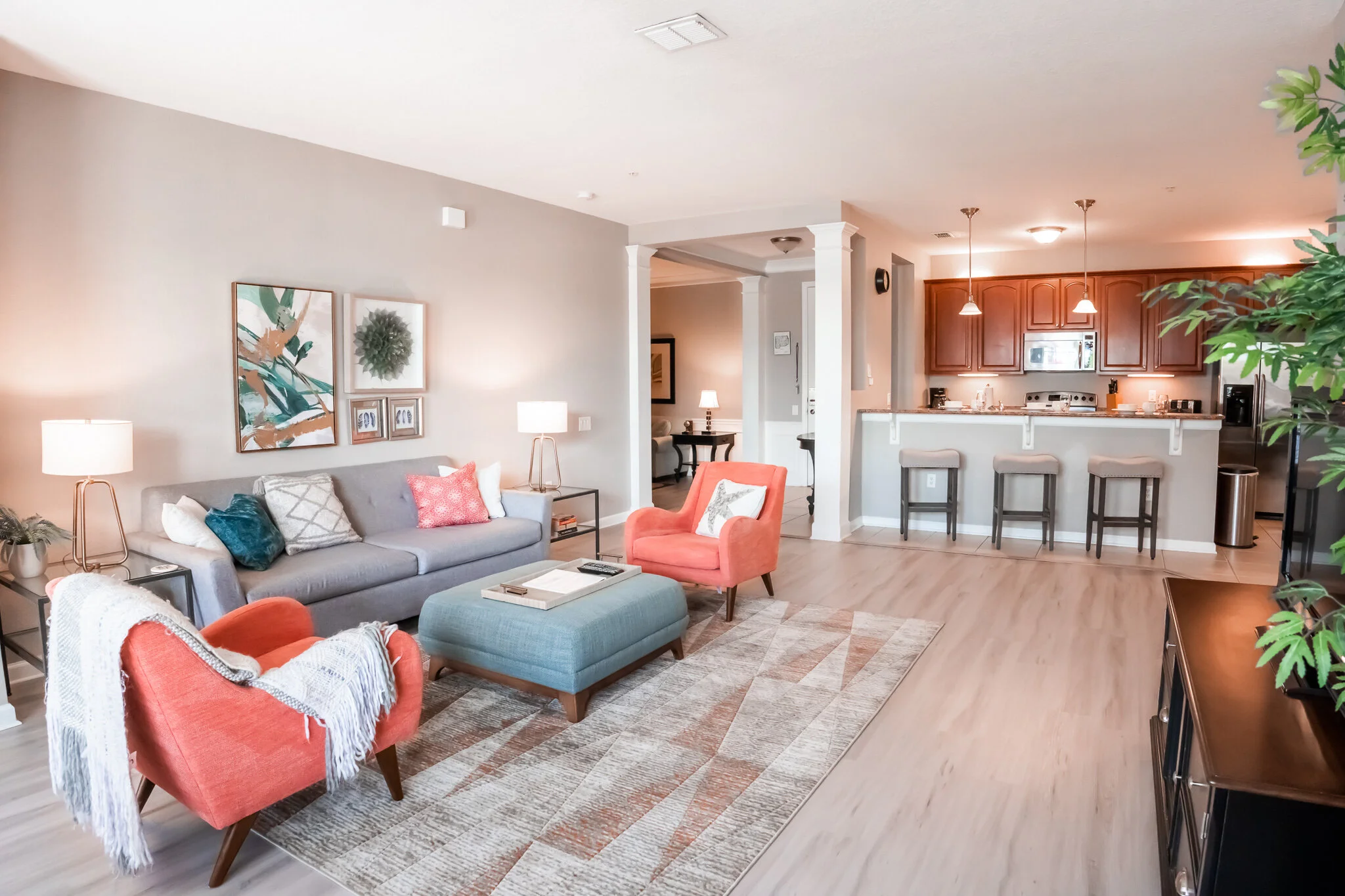 Open-concept living room and kitchen with modern furnishings, a gray sofa, coral and teal armchairs, abstract wall art, and a wooden kitchen with bar stools.