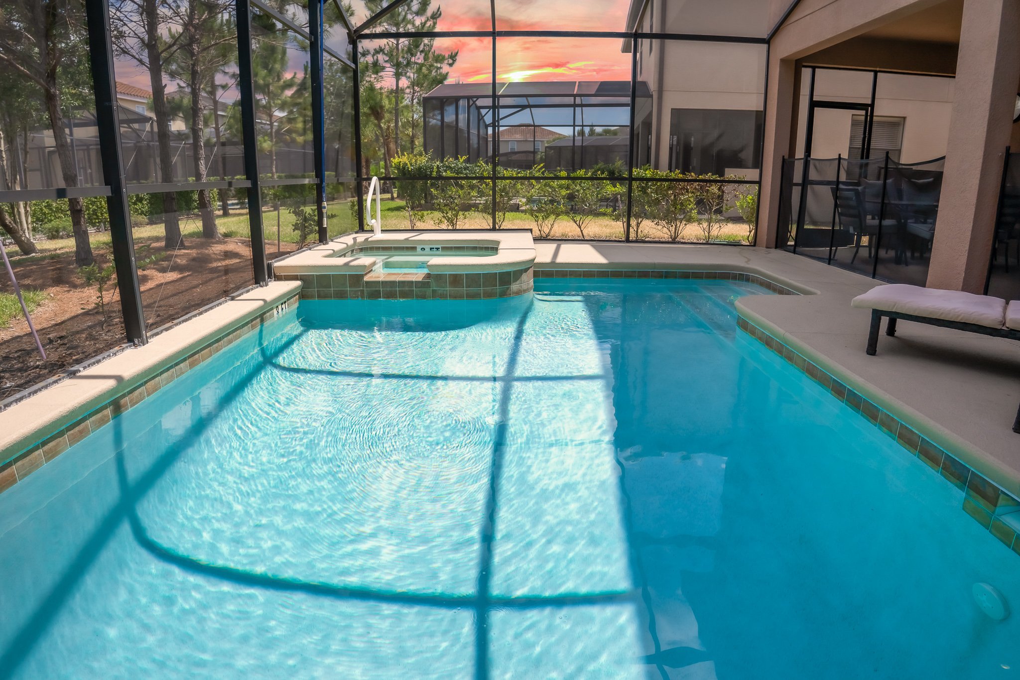 A screened-in backyard patio with a swimming pool and hot tub at sunset, surrounded by a privacy fence and trees.