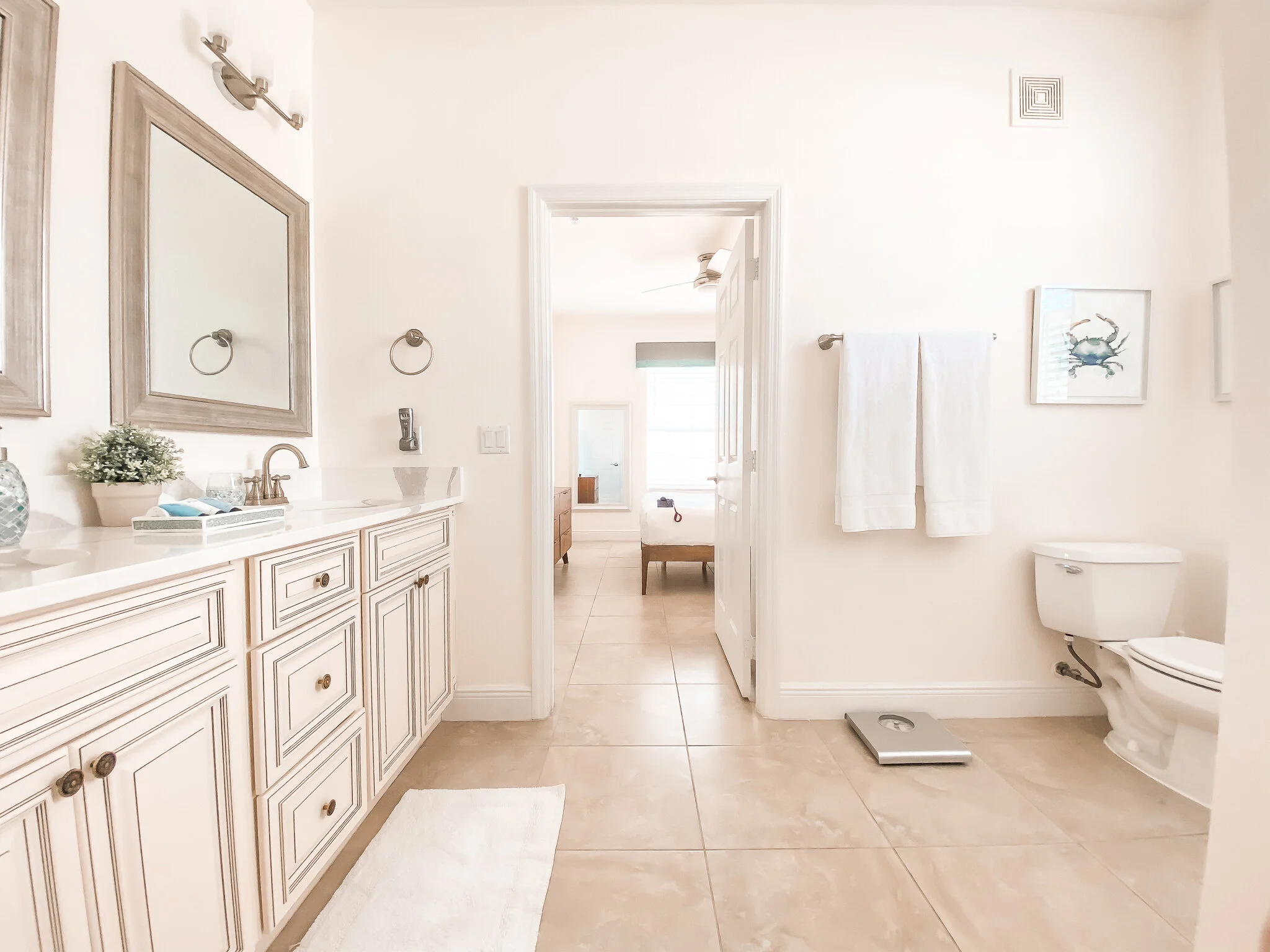 A bright, modern bathroom with a large white vanity, framed mirrors, and brass fixtures. There are white towels on a rack, a framed crab picture on the wall, and a bathroom scale on the floor. The room opens to a bedroom with a window and a bed visib