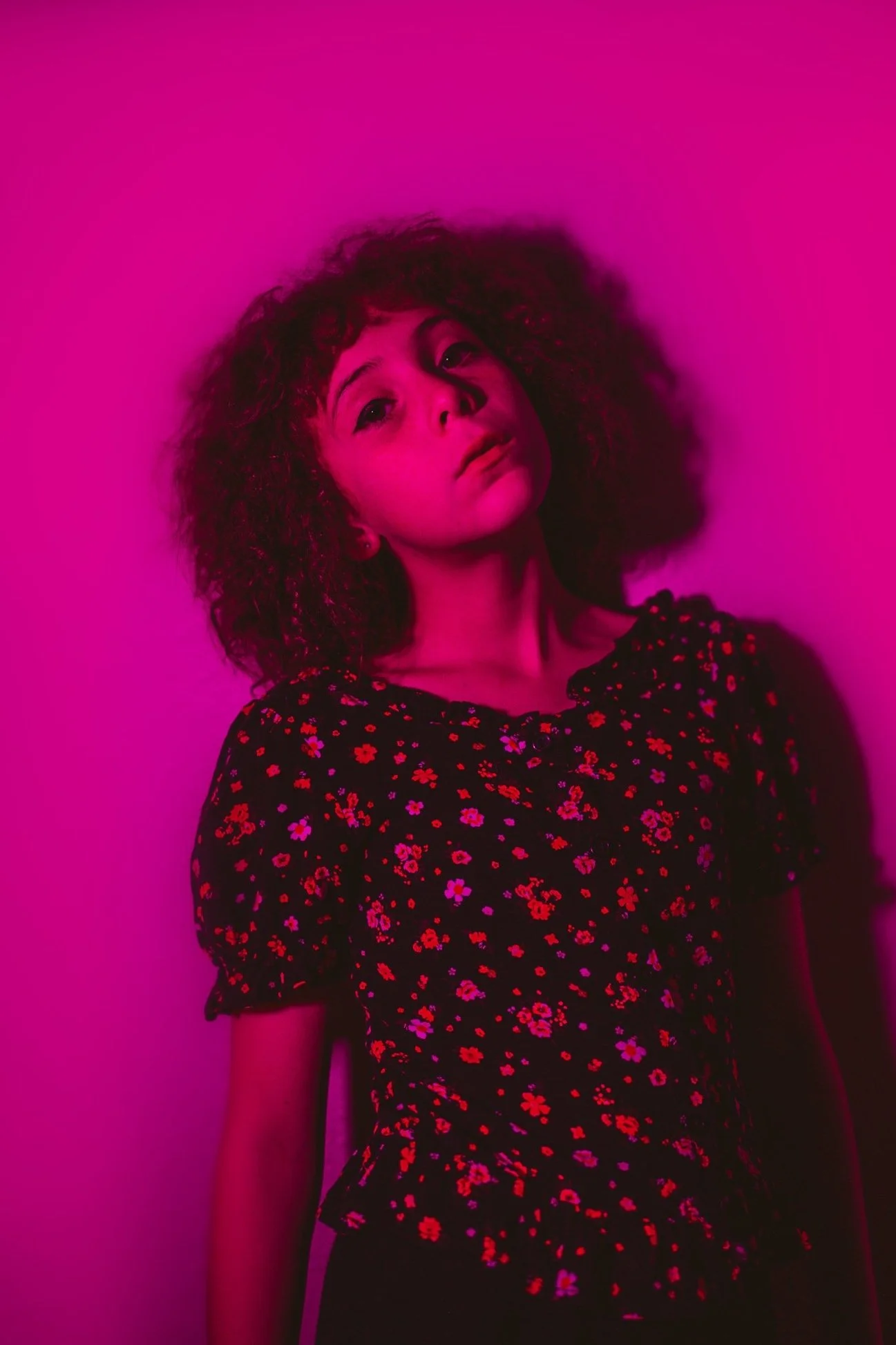 A woman with curly hair standing against a pink backdrop, wearing a black dress with a red and purple floral pattern.
