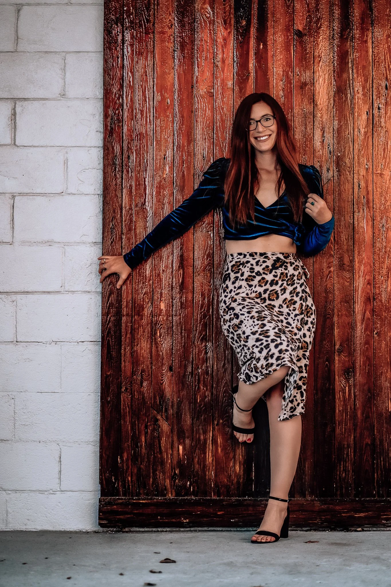 A woman with red hair and glasses, wearing a blue velvet crop top and a leopard-print skirt, standing against a wooden door with a white brick wall on the left side, smiling at the camera.