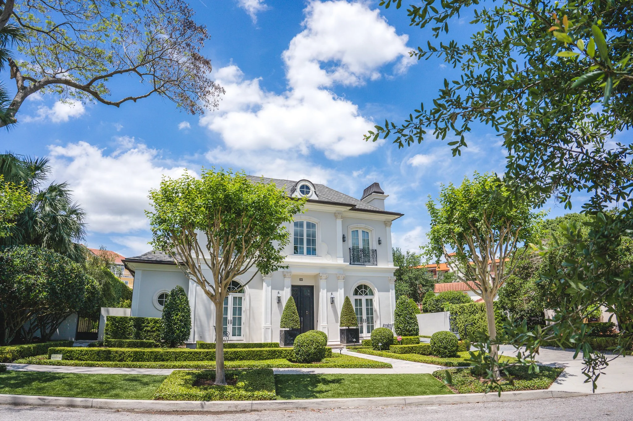 A large, white two-story house with a gray roof, surrounded by well-manicured lawn and landscaping. Tall trees with green foliage frame the house on either side. The house features arched windows, a balcony, and exterior lamps, under a bright blue sk