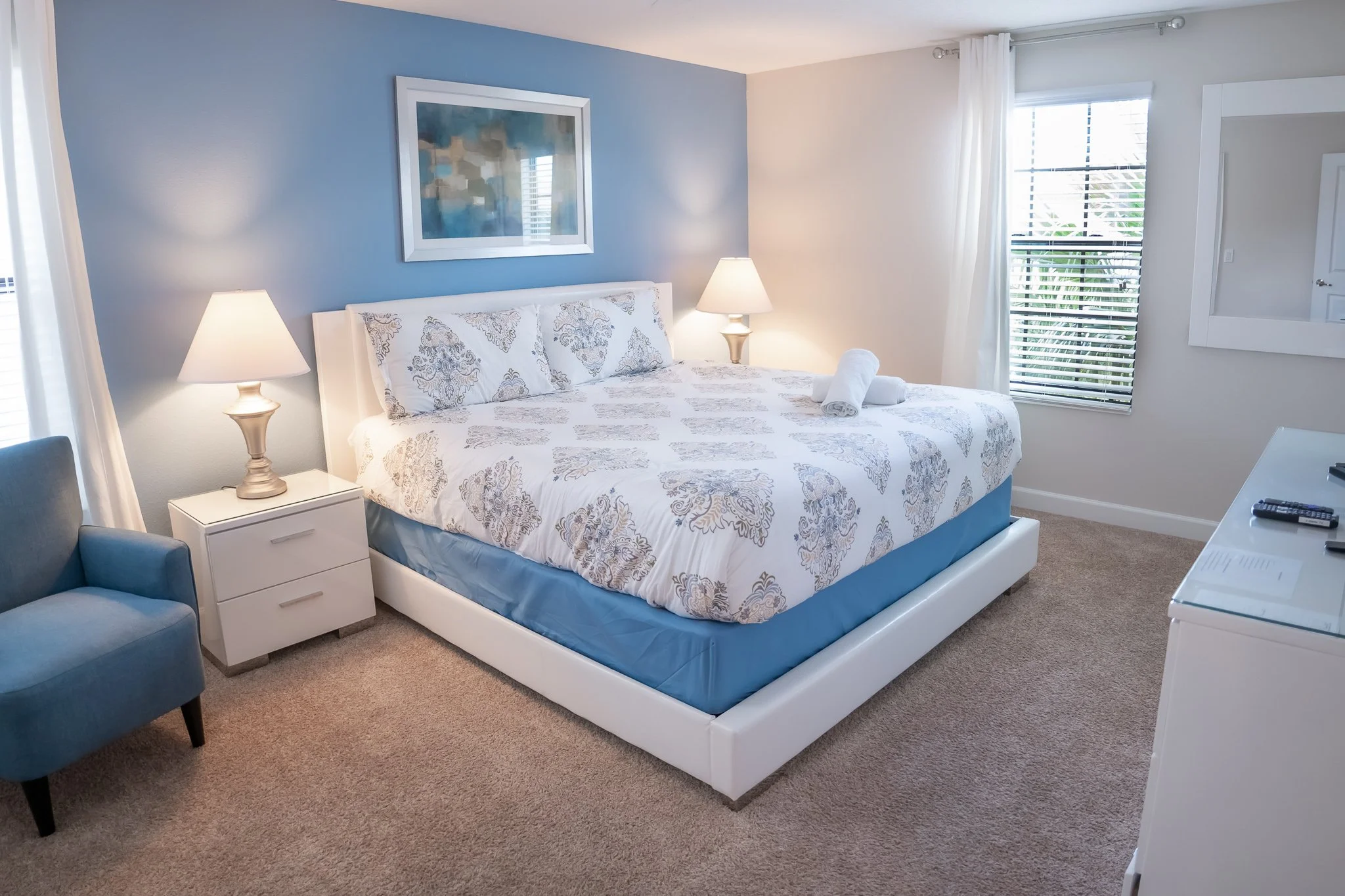 A bedroom with a neatly made bed, white and gray patterned bedding, and two rolled towels on top. The room has a blue accent wall, two lamps on white nightstands, a blue armchair, and a large window with white curtains and blinds.