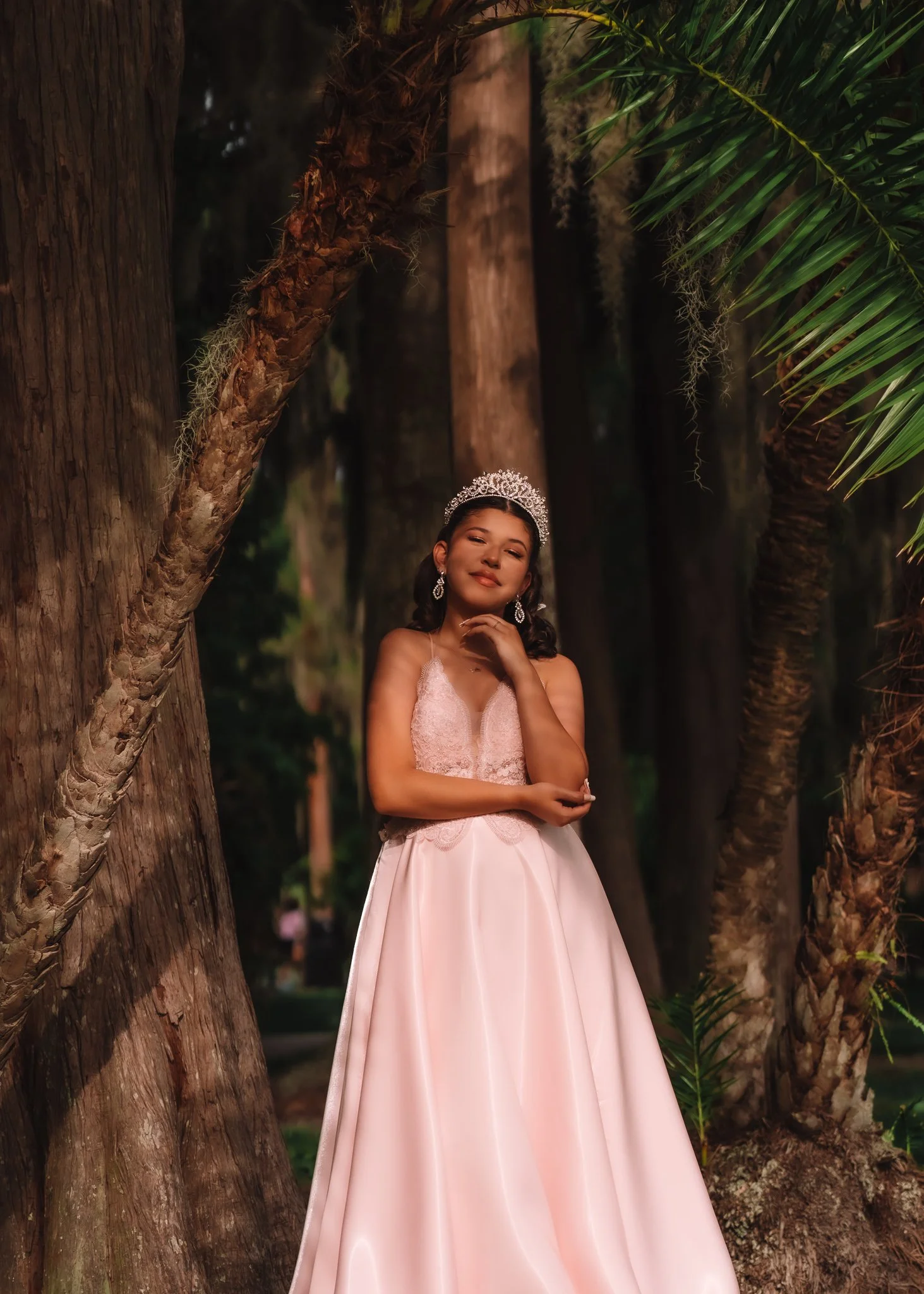 A young woman in a pink ball gown and tiara standing among tall trees with palm leaves in a forest setting.