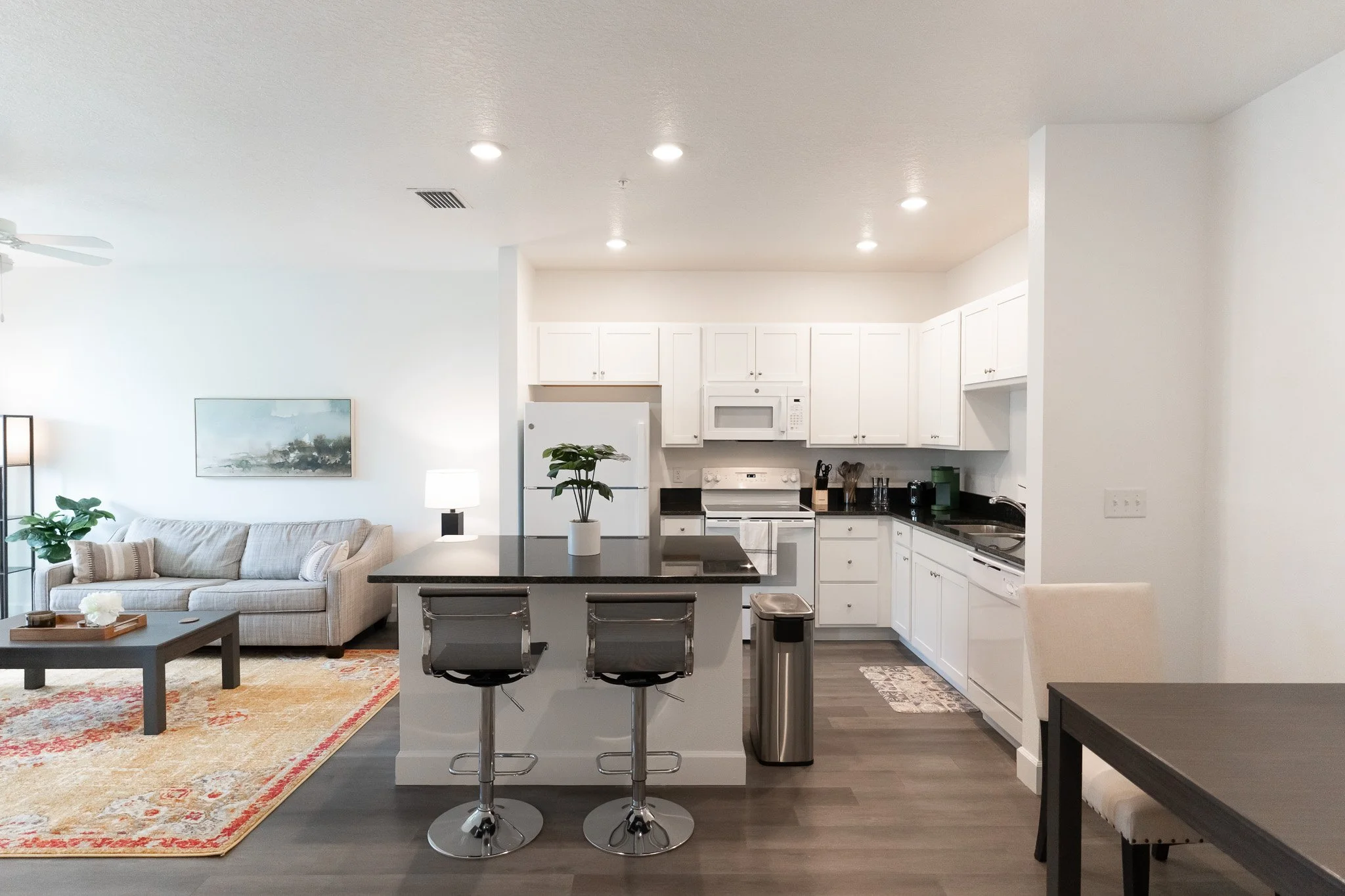 Open-concept living room and kitchen with white cabinets, black countertops, and modern furniture, including a gray sofa, black coffee table, and a dining table. The kitchen features a refrigerator, microwave, stove, and a small kitchen island with t