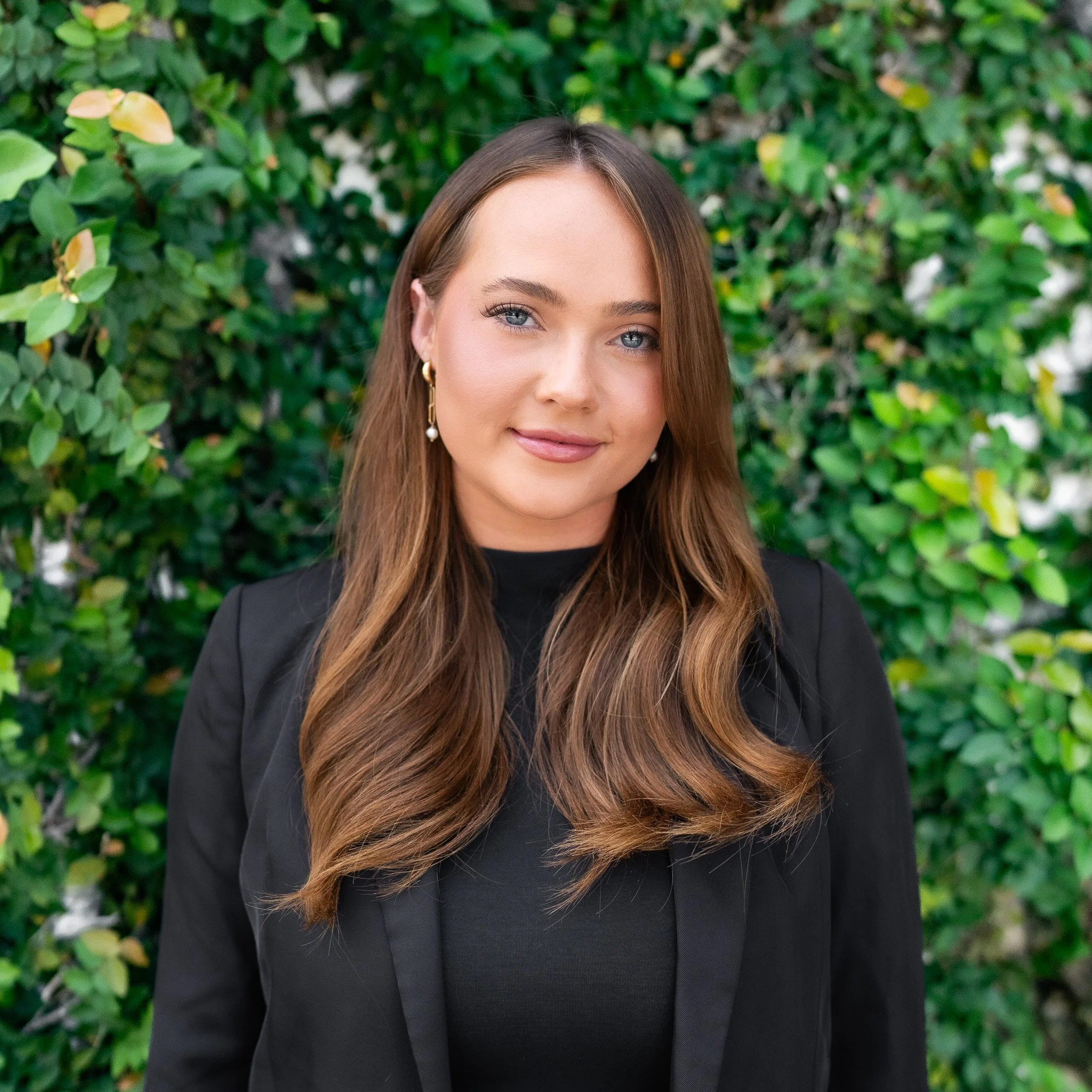A woman with long, wavy brown hair, blue eyes, and light makeup, wearing gold earrings with pearls, dressed in a black blazer and black top, standing outdoors in front of a lush green leafy background.