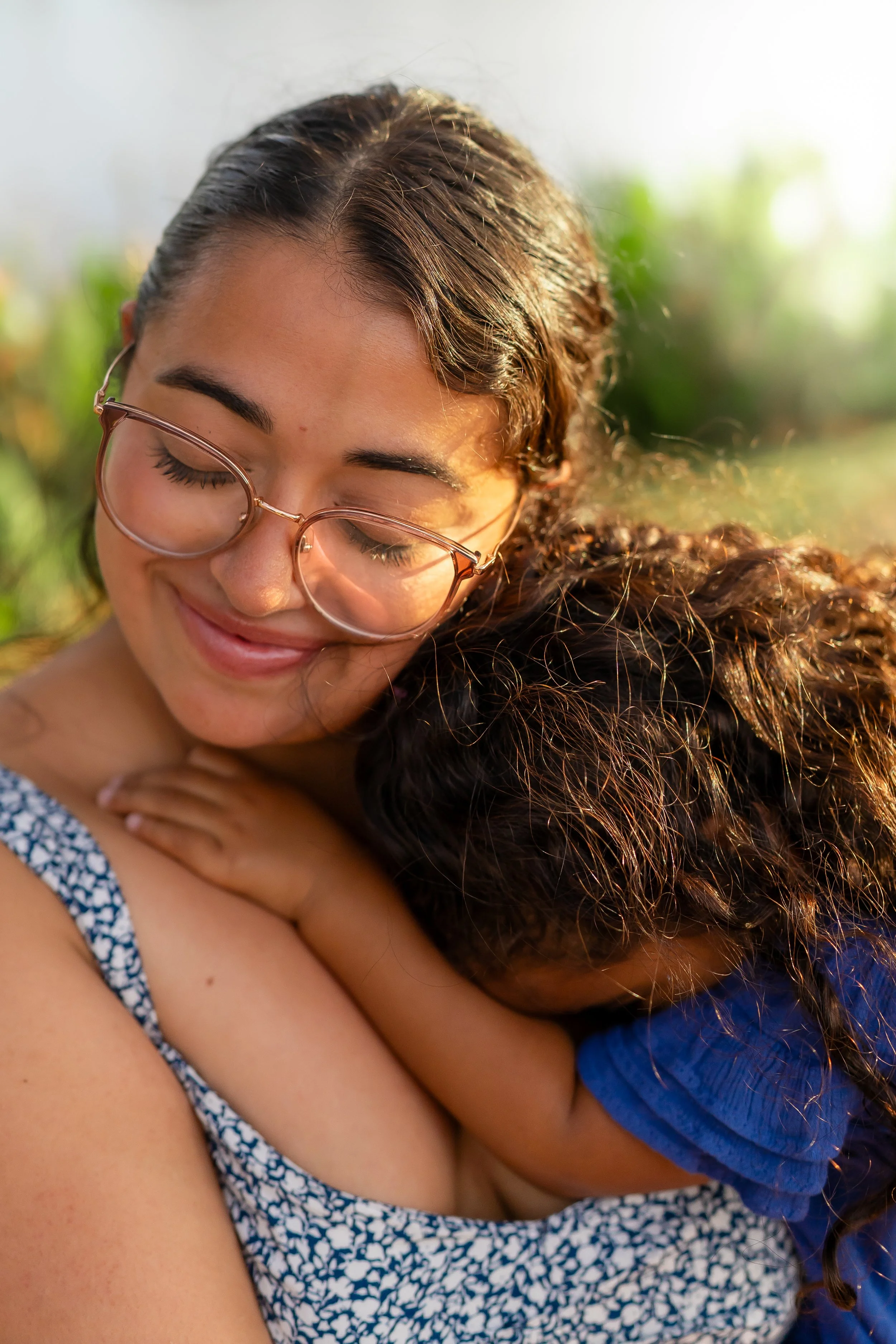 A woman with glasses smiling and hugging a child with curly hair outdoors.