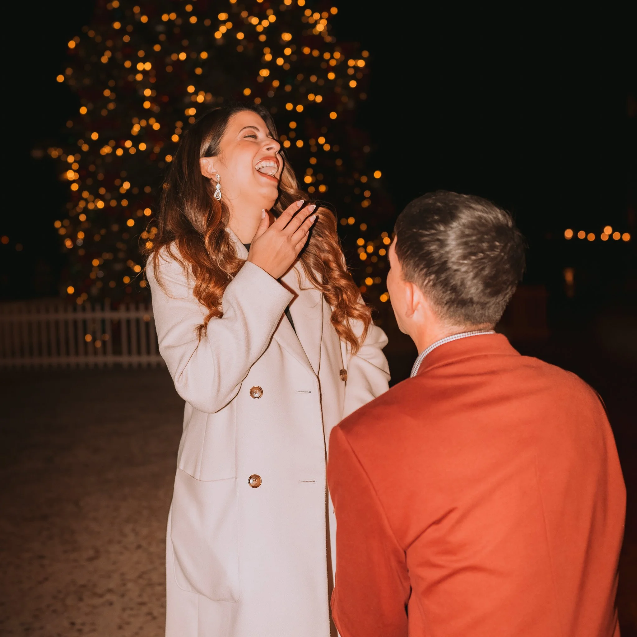 A woman with long, wavy hair and earrings laughs joyfully while holding her hand to her chest. She is dressed in a light-colored blazer and a black top. A man with short hair and a reddish-brown jacket faces her. They are outdoors at night with a lar
