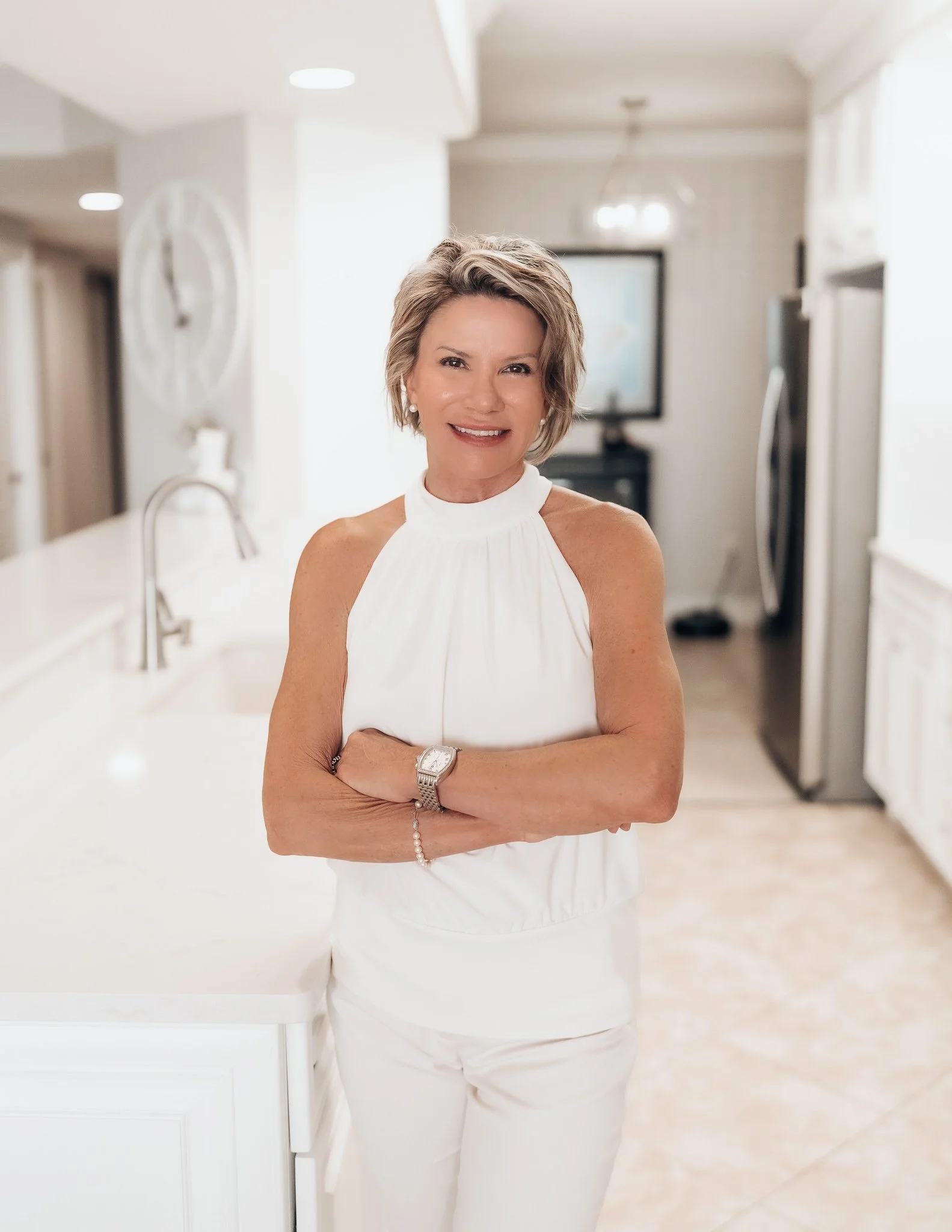 A smiling middle-aged woman with short gray hair, wearing a white high-neck sleeveless top, standing in a bright, modern kitchen with white cabinets, a stainless steel sink, and a flat-screen TV in the background.