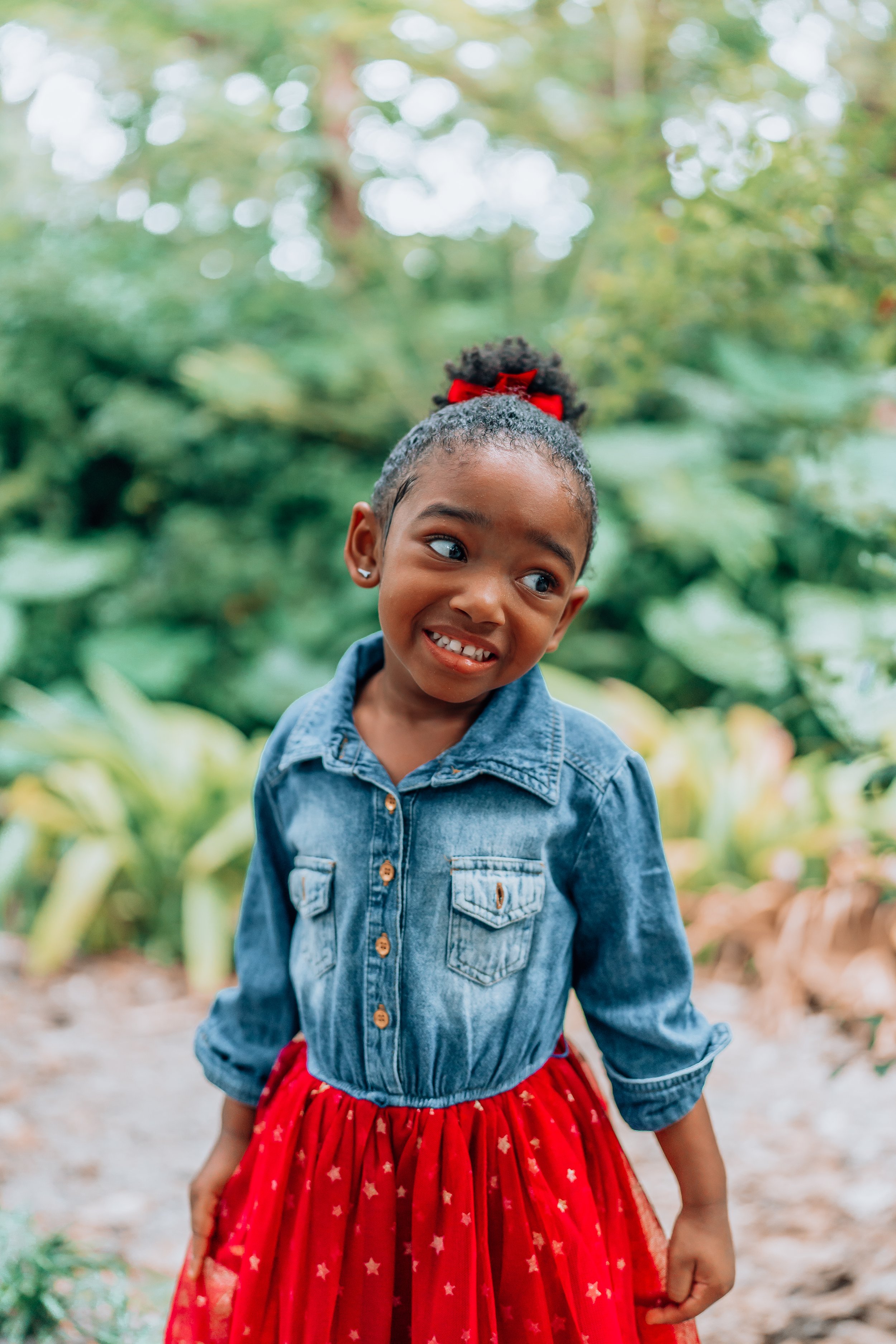 A young girl with curly hair in a puff tied with a red bow, wearing earrings, a denim jacket, and a red skirt with star patterns, standing outdoors among greenery, making a silly face.