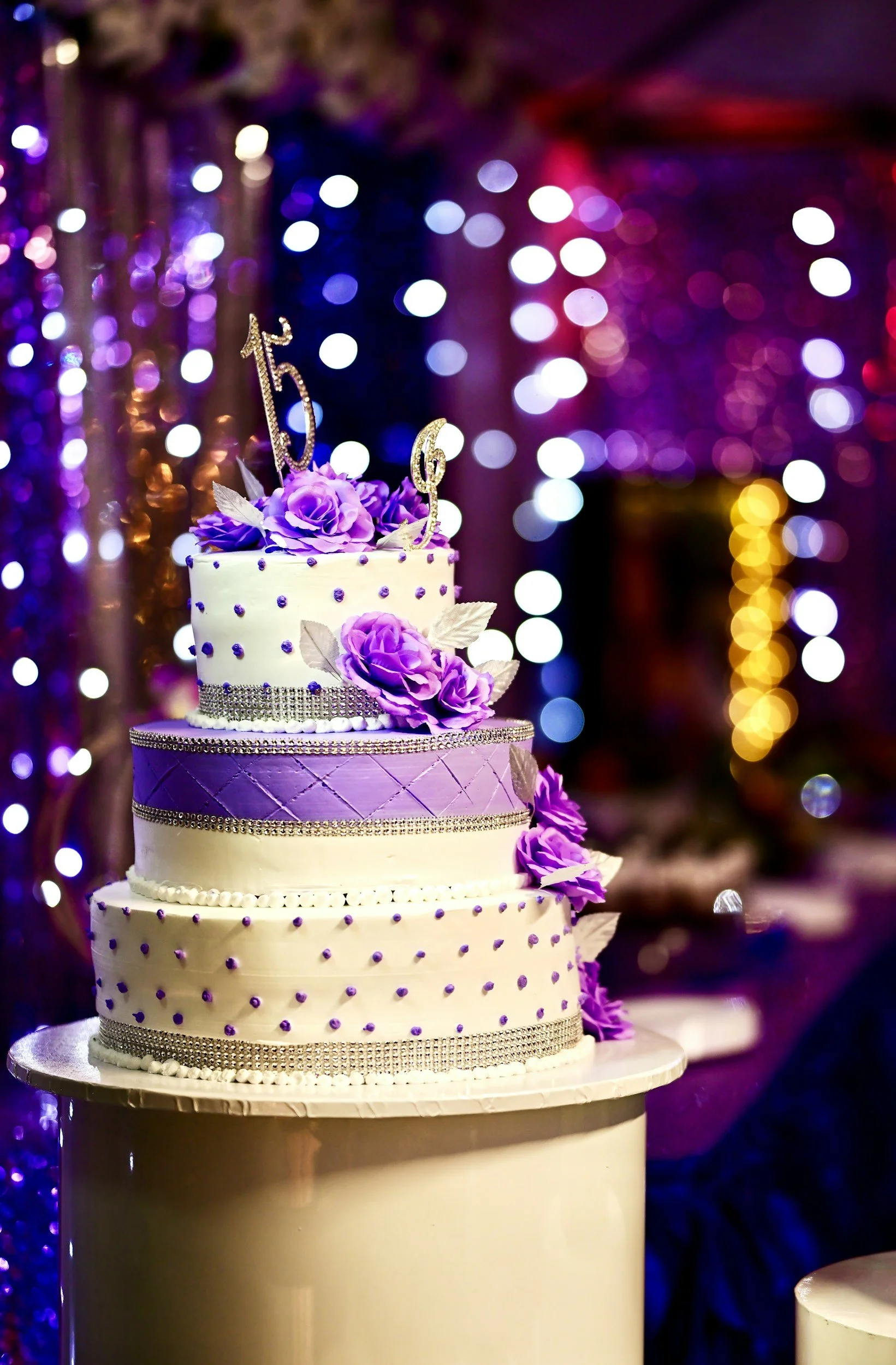 A four-tiered wedding cake decorated with purple and white flowers, rhinestone accents, and a "15" cake topper with purple roses, set against a colorful blurred background of lights and decorations.