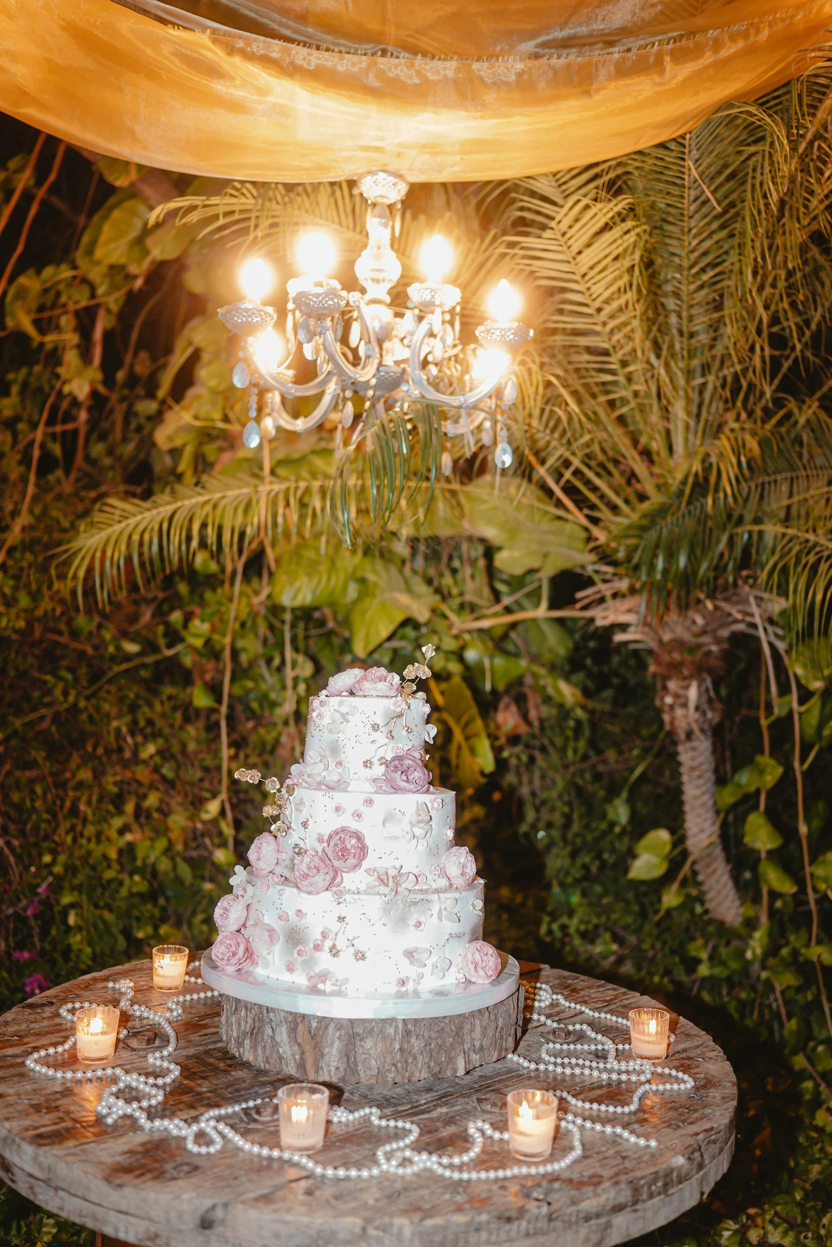 A three-tier white wedding cake decorated with pink flowers and metallic accents, placed on a wooden log slice, surrounded by candles and pearl necklaces on a rustic table, with lush green foliage and a chandelier hanging above.