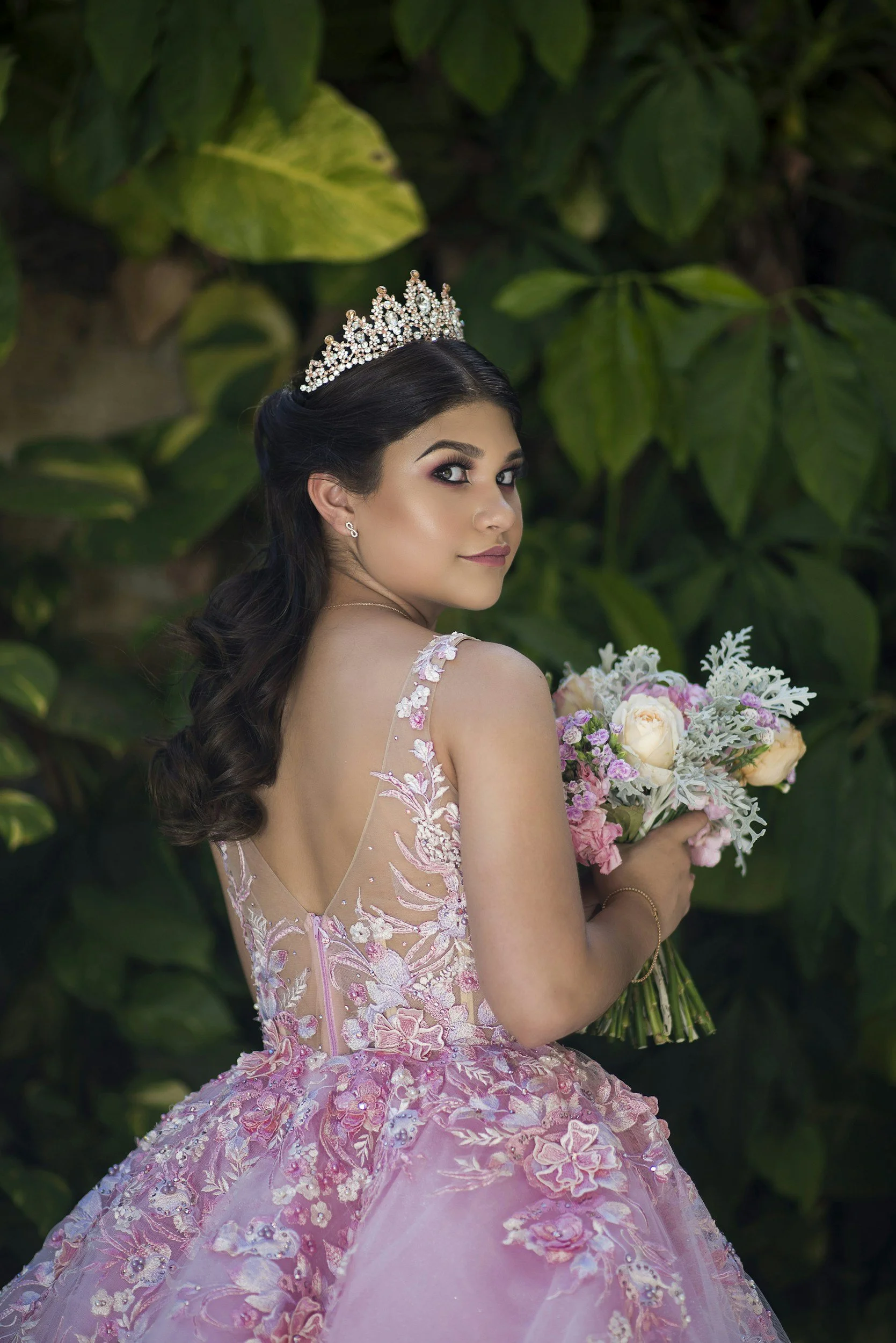 A young woman in a pink floral gown holding a bouquet of flowers. She is wearing a tiara and earrings, and has dark, wavy hair styled, with a background of large green leaves.