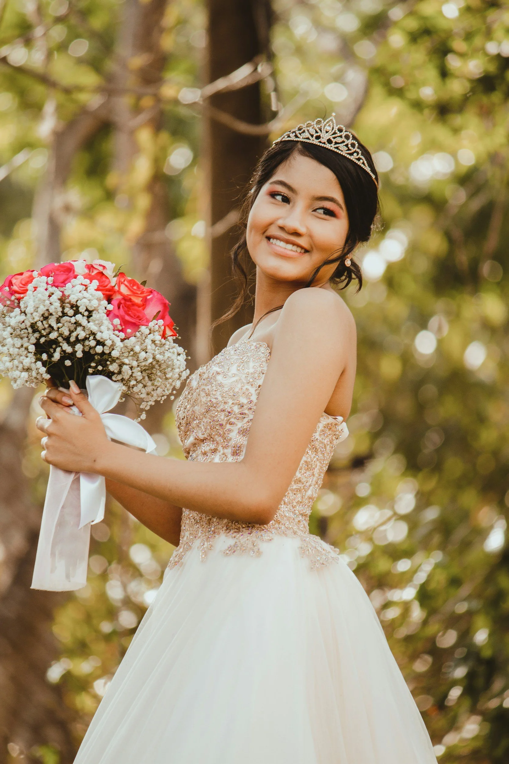 A young woman wearing a tiara and a strapless gown, holding a bouquet of pink roses and baby's breath, standing outdoors with a blurred background of trees and sunlight.