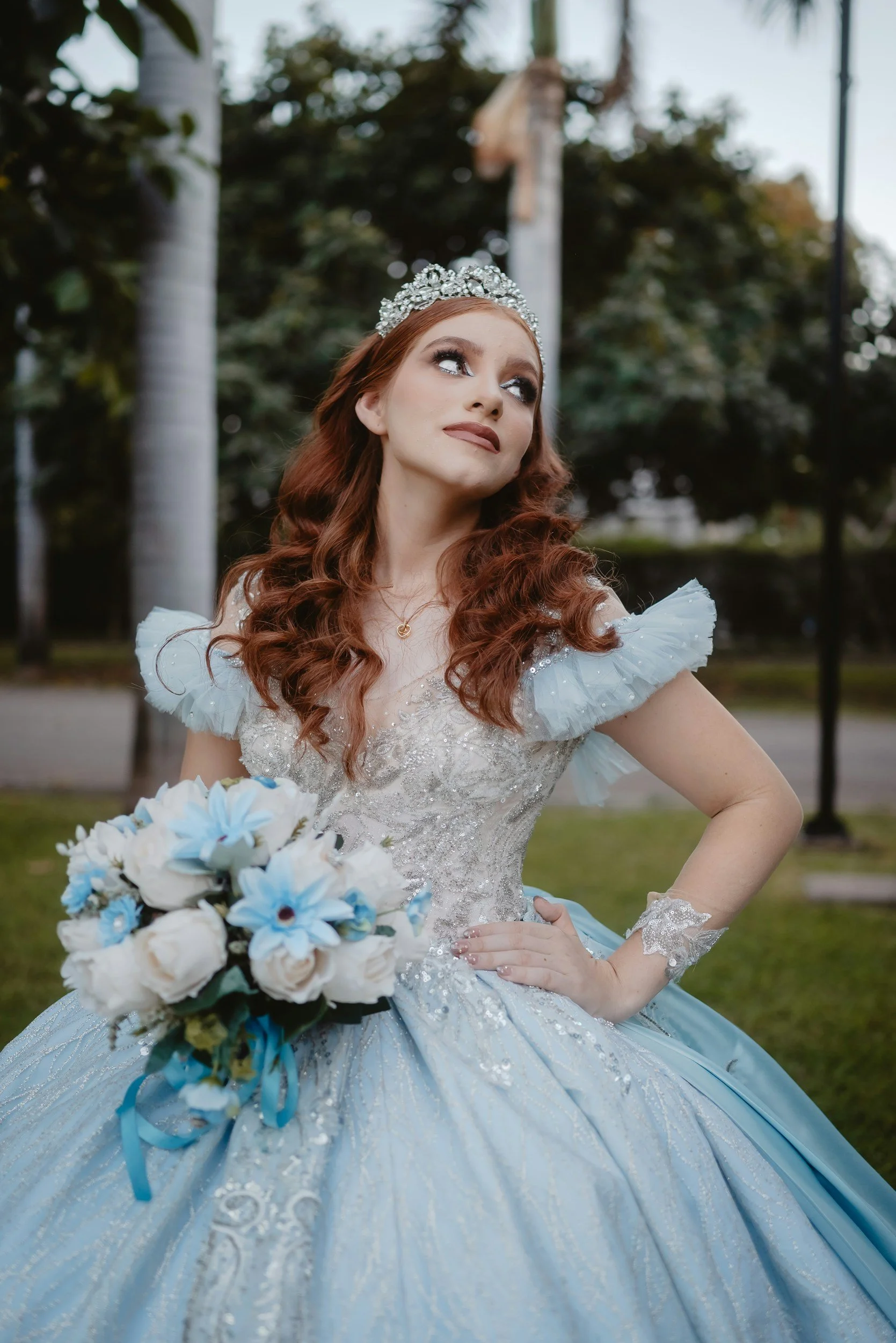 A woman with red hair wearing a tiara and a wedding dress holding a bouquet of white and blue flowers outdoors.