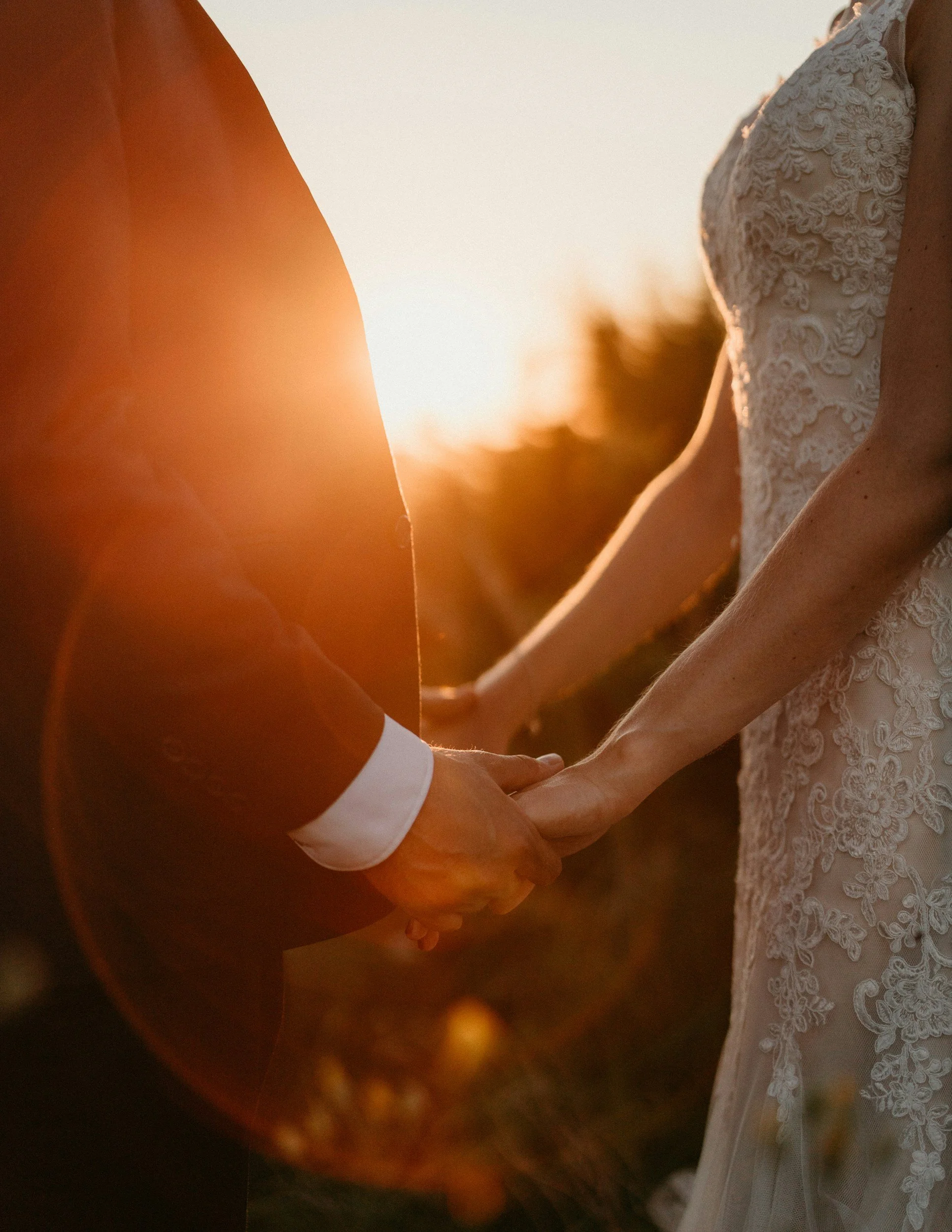 A man and woman holding hands during sunset, with the woman wearing a wedding dress with lace details.
