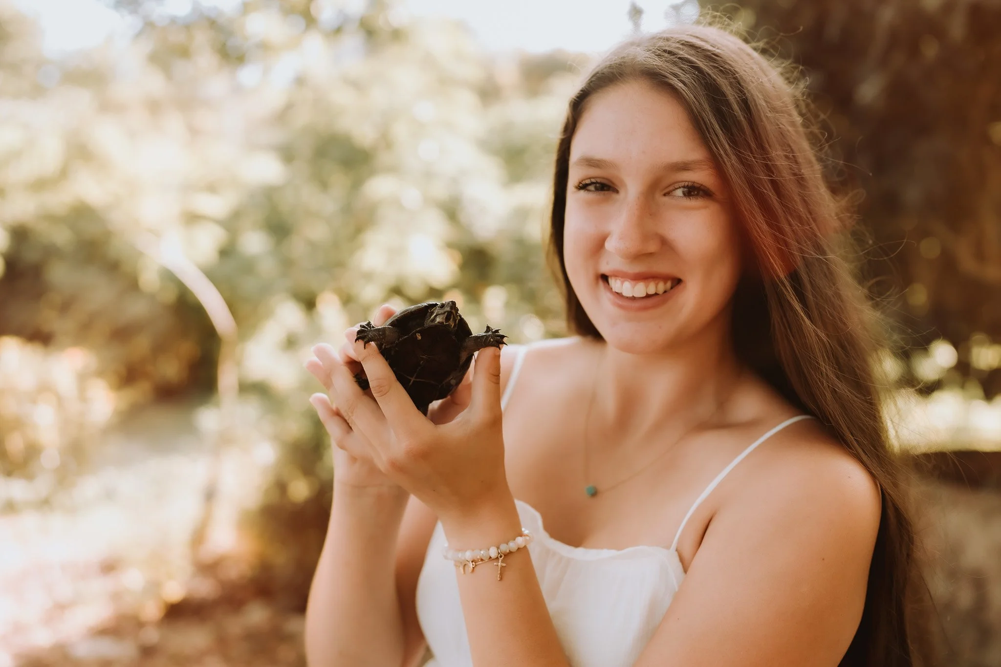 A young woman with long brown hair smiling outdoors while holding a small turtle in her hands