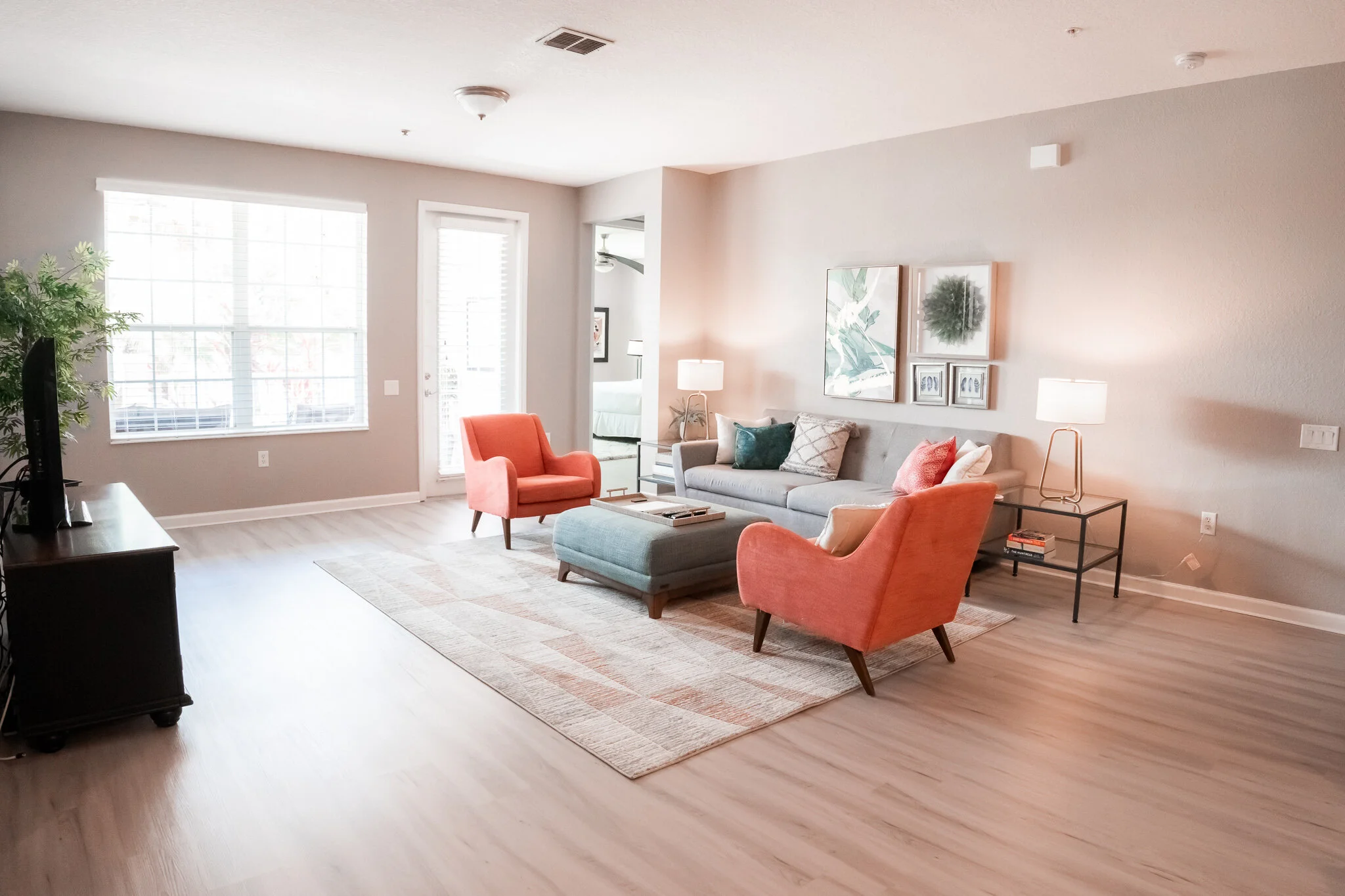 Bright living room with light-colored hardwood floors, a large window, a gray couch with pillows, two coral-colored armchairs, a gray ottoman, a black side table with a lamp, and framed artwork on the wall.