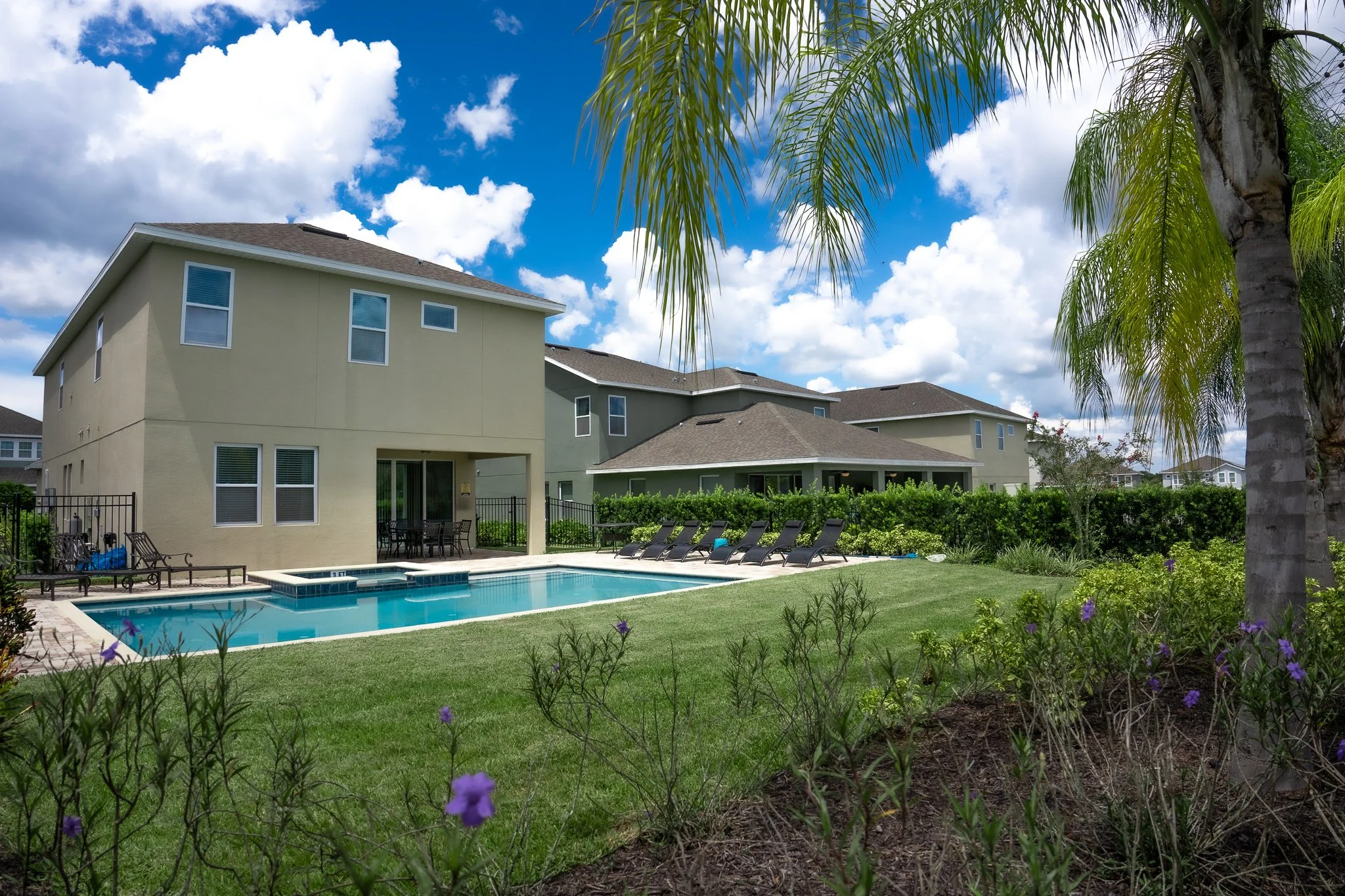 A backyard with a swimming pool, surrounded by lounge chairs, greenery, and houses under a bright blue sky with clouds.