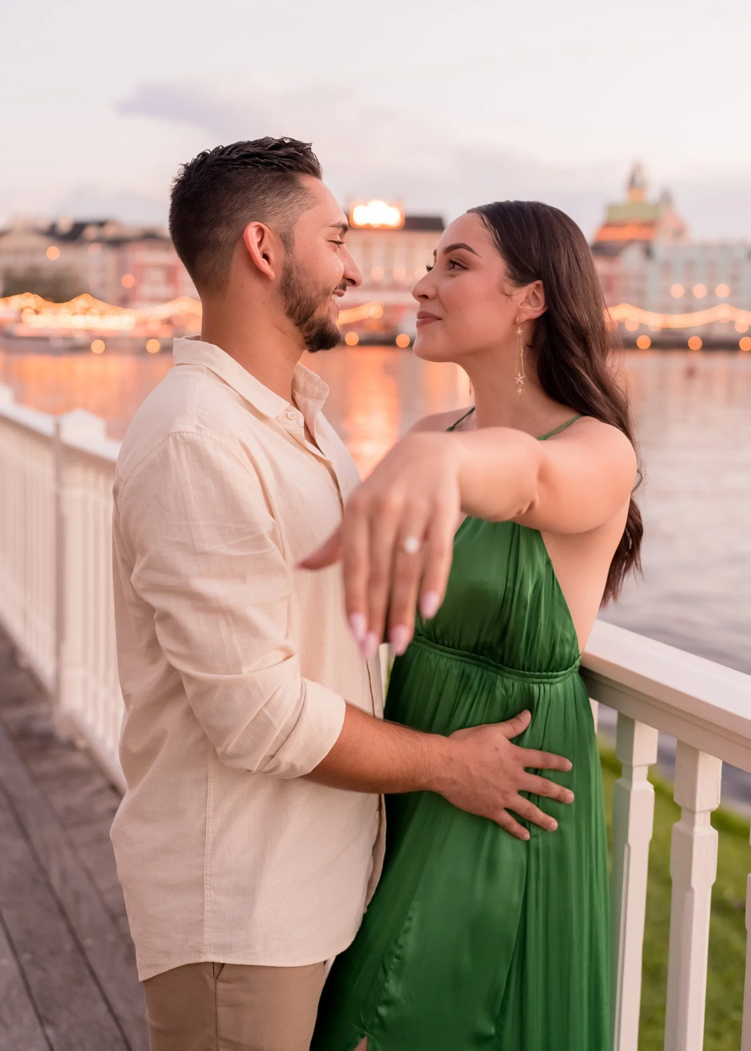 A couple is dancing closely by a river at sunset, with city buildings and lights in the background. The man wears a light-colored shirt and the woman wears a green dress.