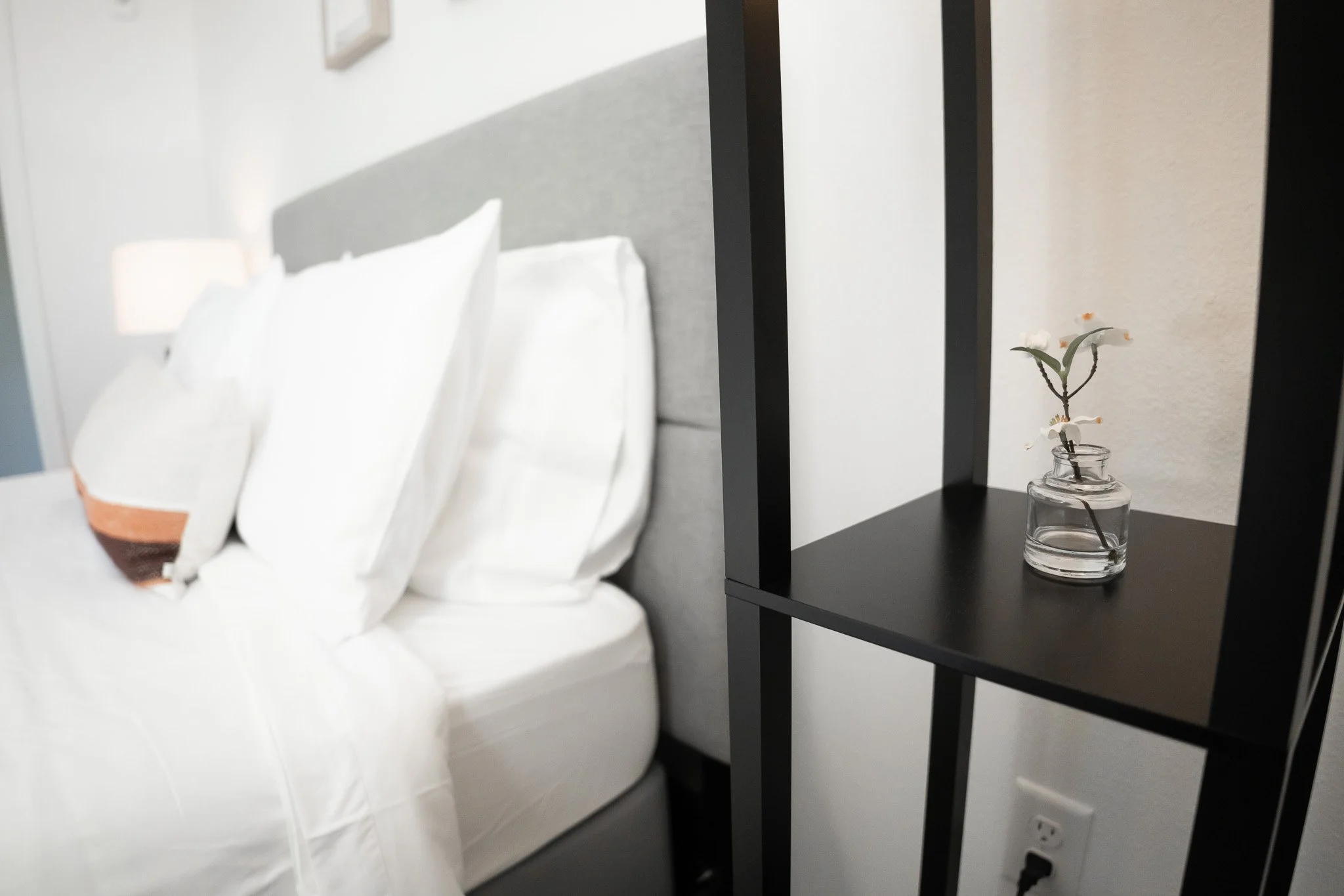 Close-up of a black shelving unit with a small glass vase containing a white flower on a bedside table in a bedroom with white bedding and a gray headboard.