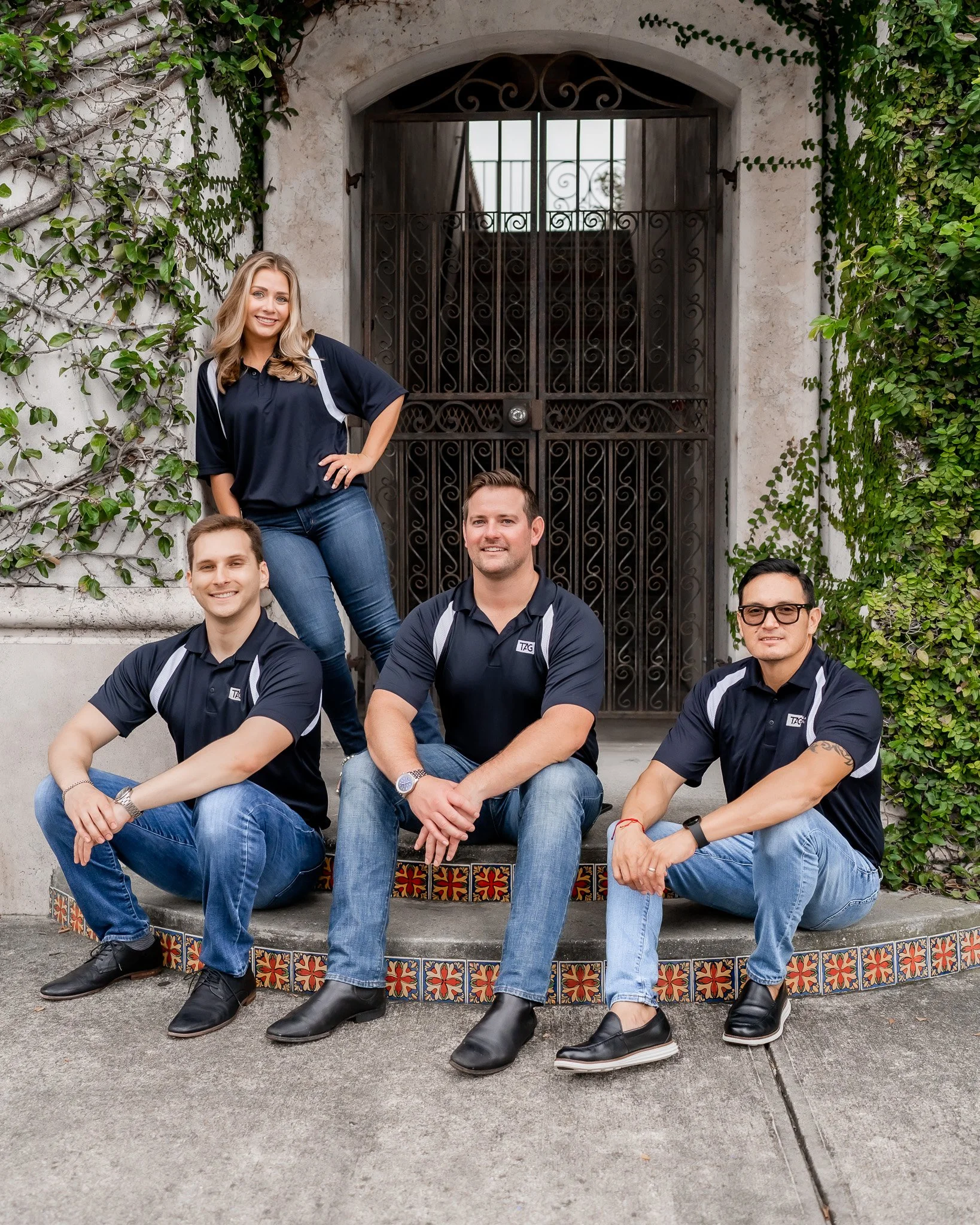 Four people wearing matching navy blue and gray shirts, three men sitting on a colorful tiled step and one woman standing behind them, in front of a black iron gate surrounded by green ivy.