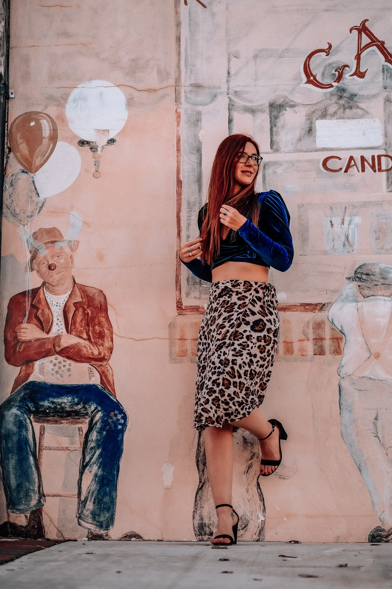 A woman with long red hair and glasses wearing a blue velvet long sleeve crop top, a leopard print skirt, and black high heels posing against a wall with painted murals of a seated bear holding balloons and a duck.