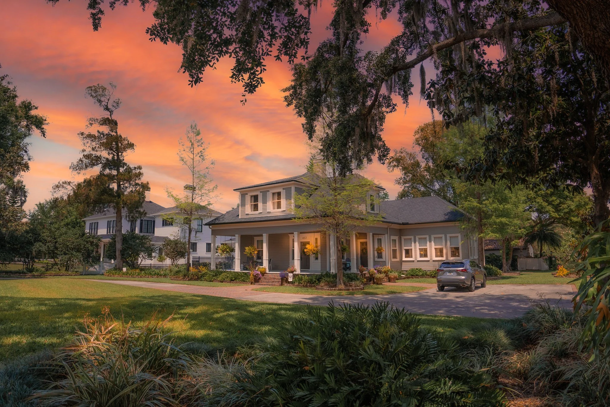 A large house with a porch decorated with hanging plants and flowers, set on a well-manicured lawn at sunset. There are cars parked in the driveway, and other houses and tall trees are visible in the background.