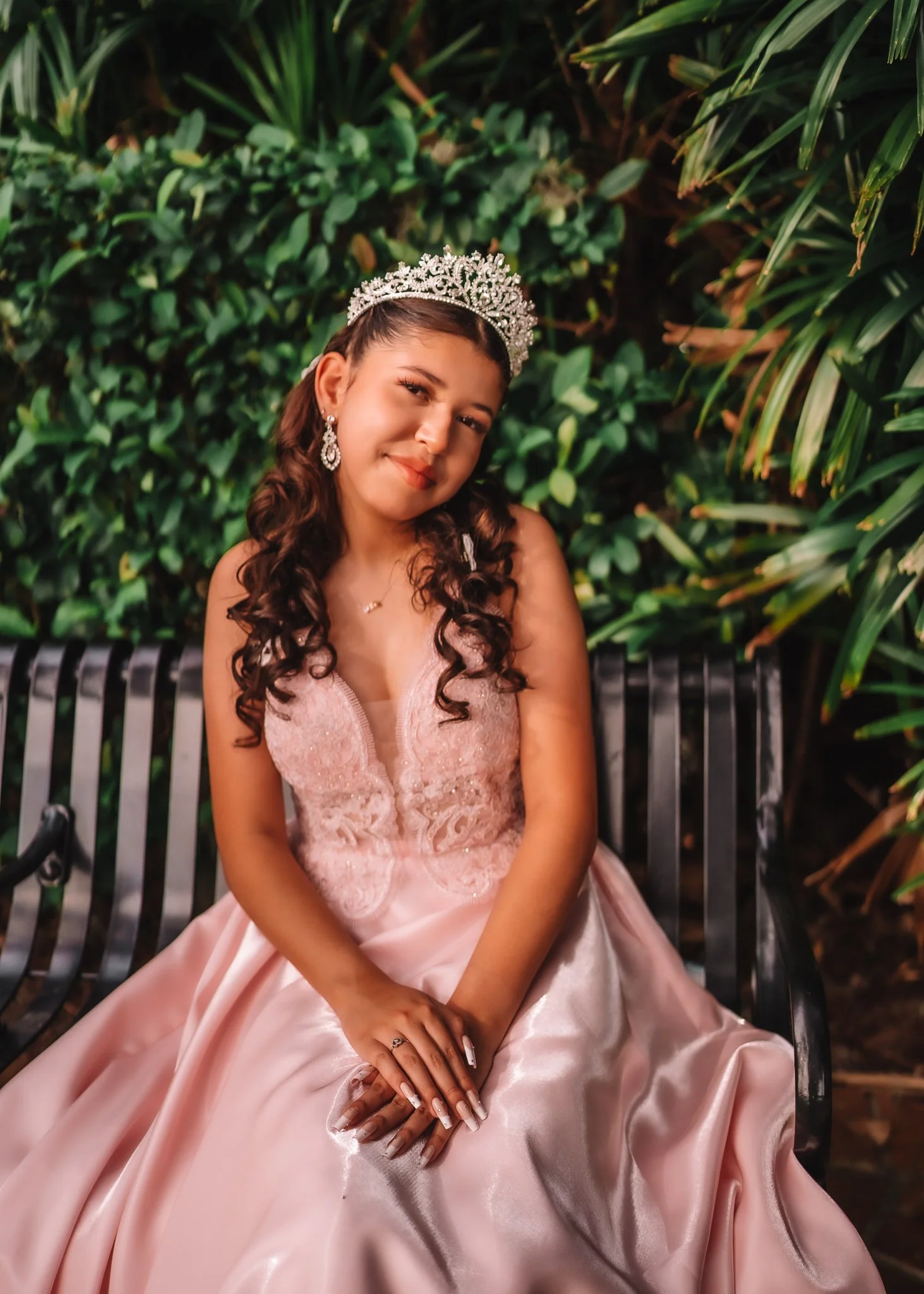 A young woman in a pink dress and tiara sitting on a black park bench with green foliage background, smiling softly.