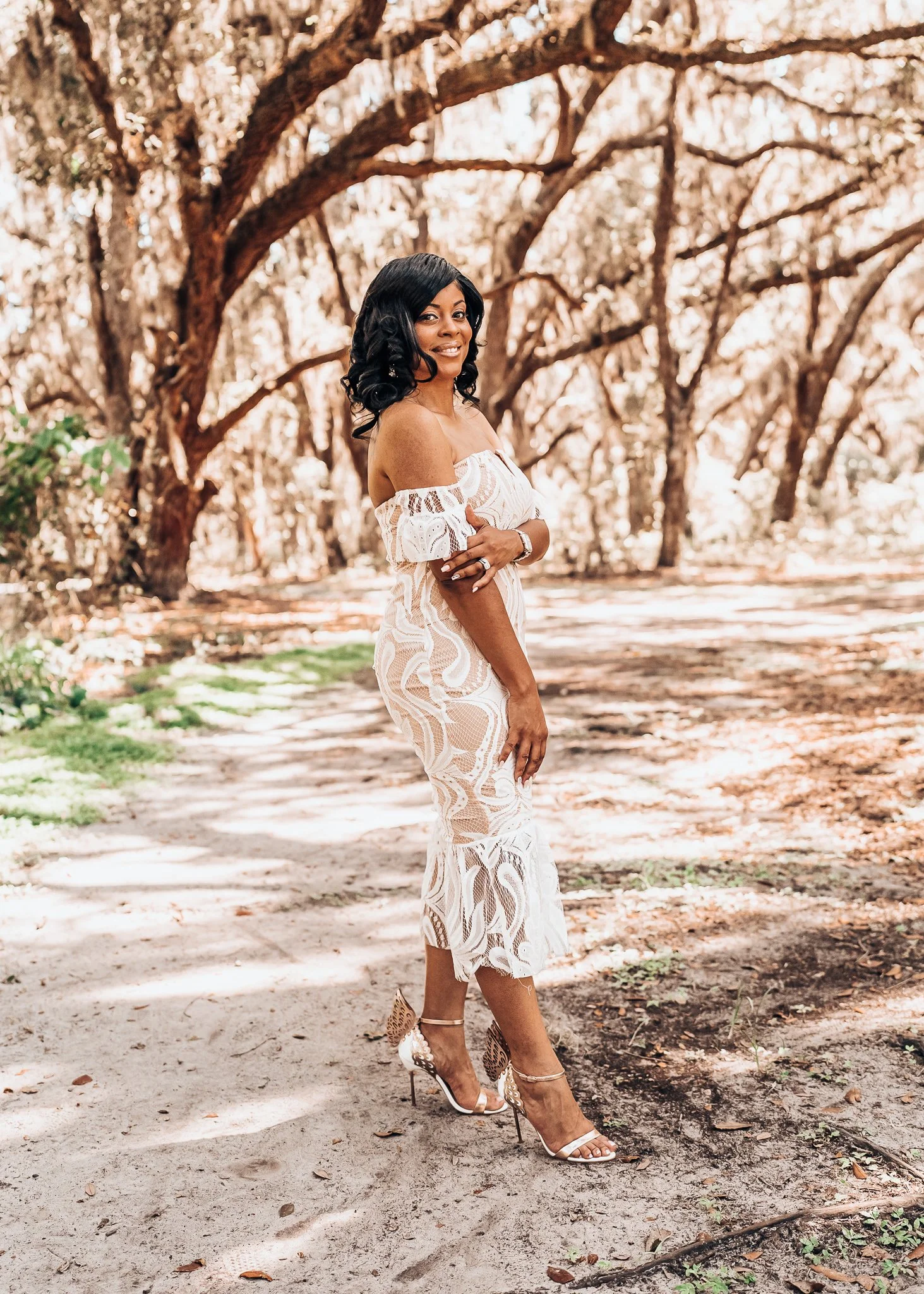 Woman in a white lace off-shoulder dress and high heels standing outdoors on a sandy path, surrounded by trees with twisted branches.