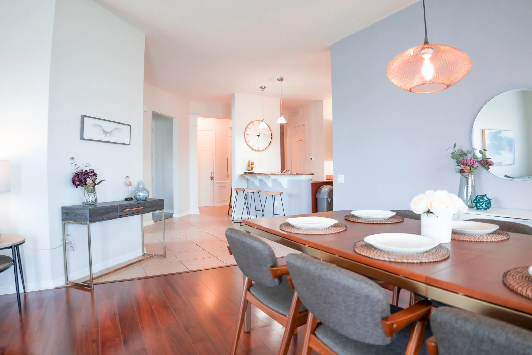 Modern dining room with a wooden table set for six, featuring white plates and woven placemats, with a flower arrangement in the center. The room has gray upholstered chairs with wooden arms, a blue accent wall with a round mirror, and a pink pendant