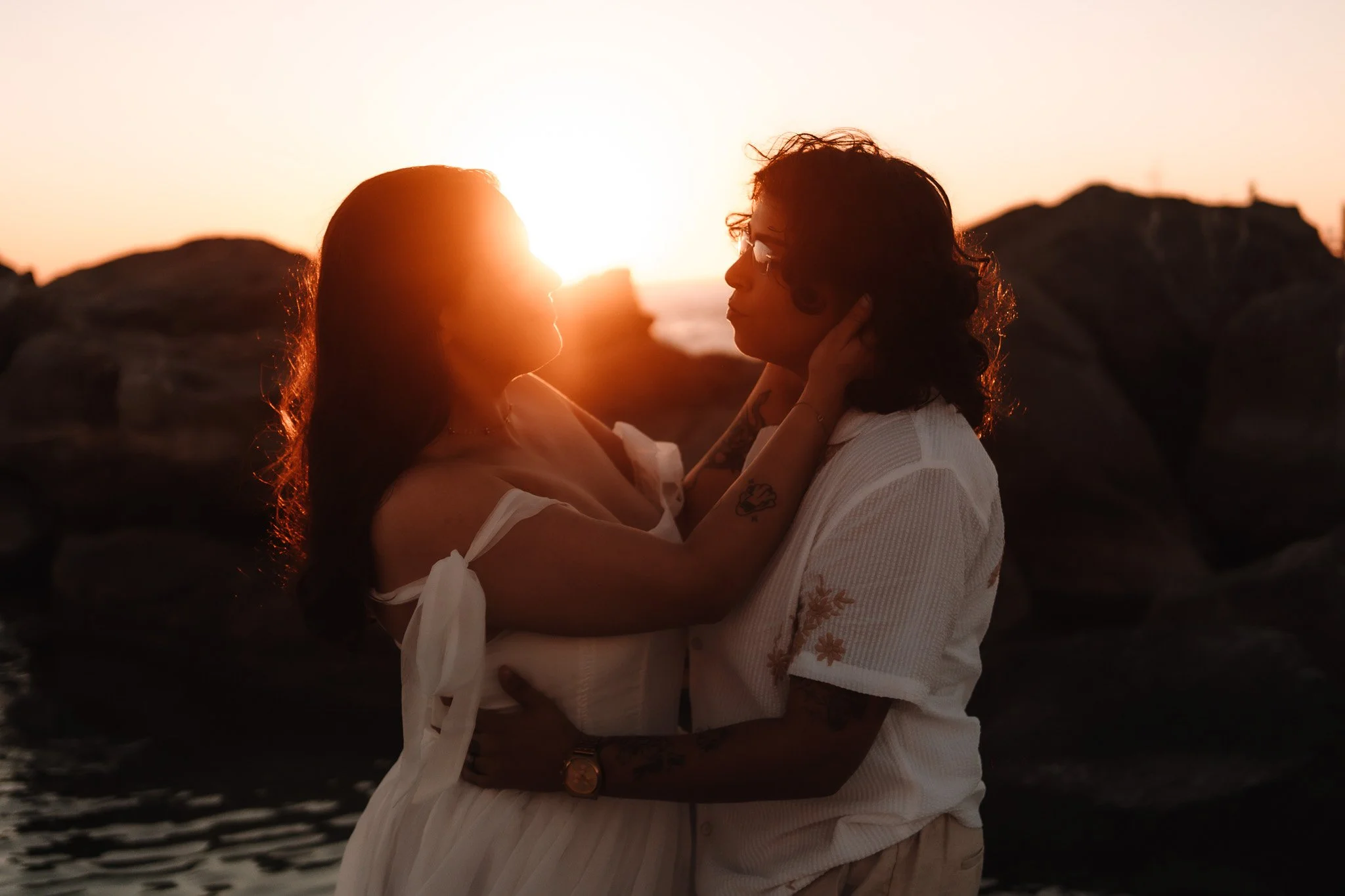 Two women facing each other, holding each other's faces, with the sunset in the background on a rocky beach.