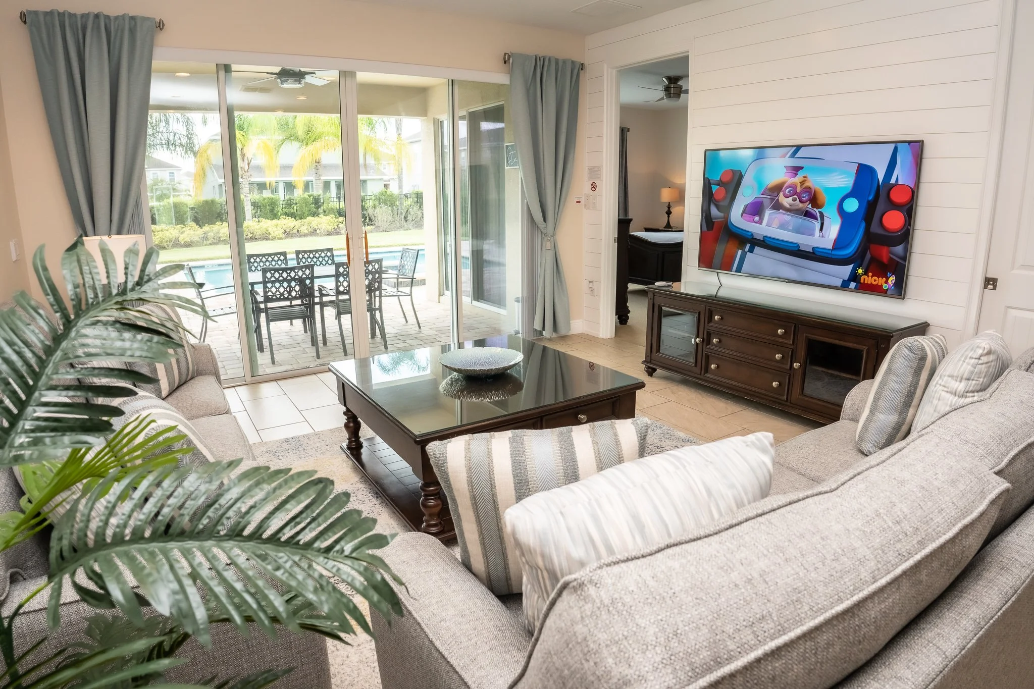 Living room with sofas, a coffee table, a TV showing a cartoon, and sliding glass doors leading to a patio and pool area.