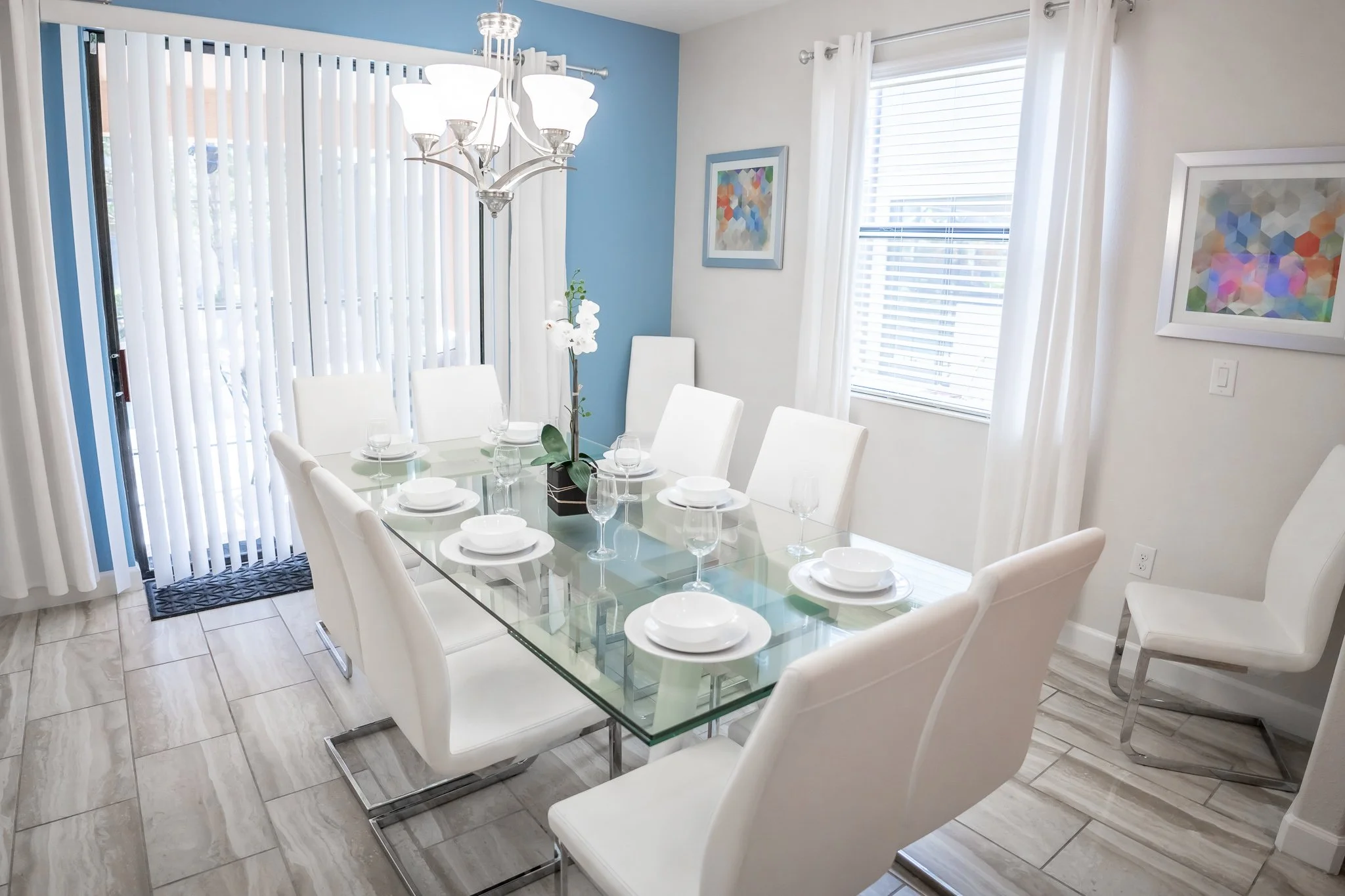 Modern dining room with a glass table, white chairs, a chandelier, and colorful artwork on the walls, with windows fitted with blinds and curtains.