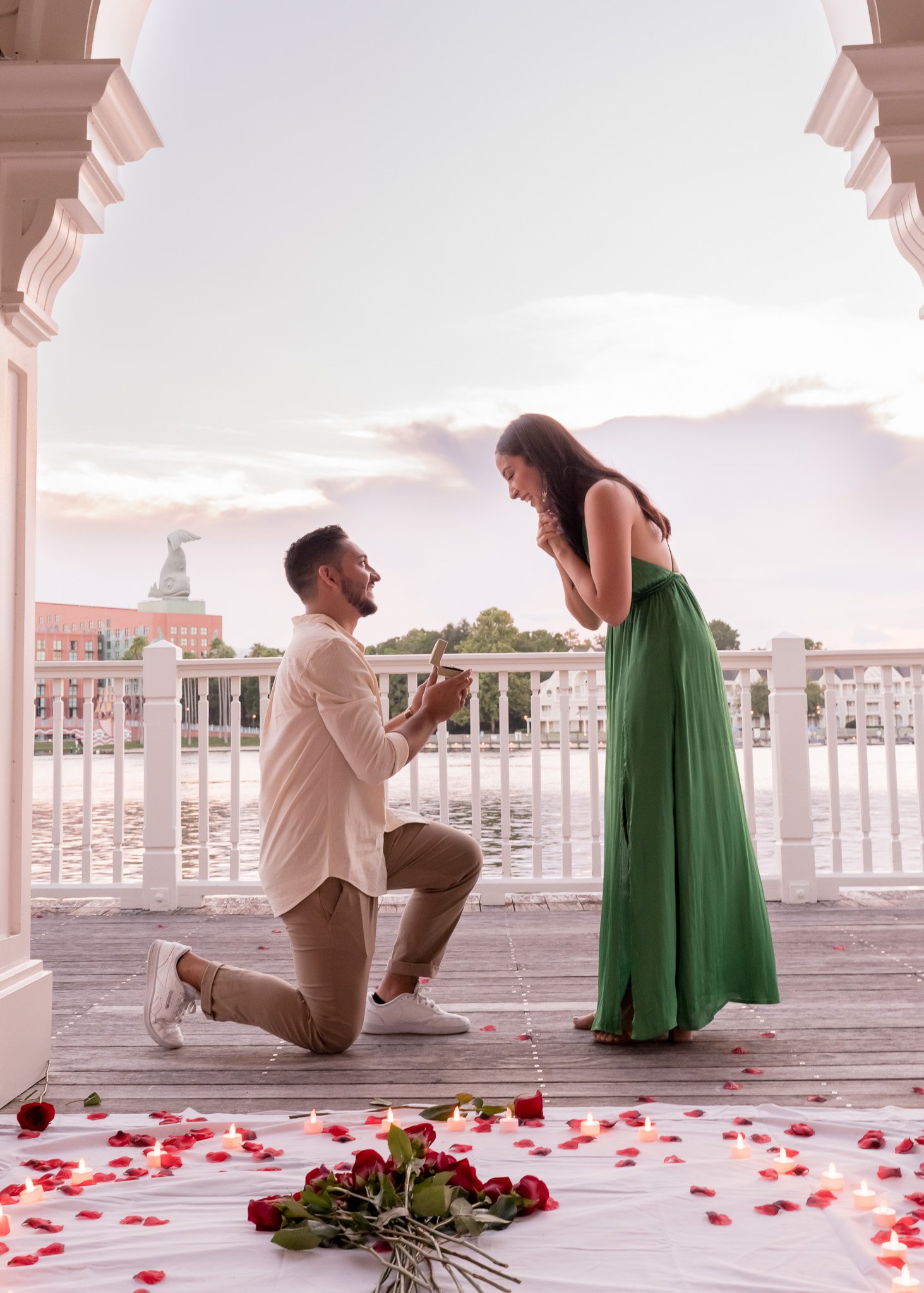 A man proposing to a woman on a romantic outdoor balcony decorated with candles, rose petals, and a bouquet of roses, with a cityscape and a large sculpture in the background at sunset.
