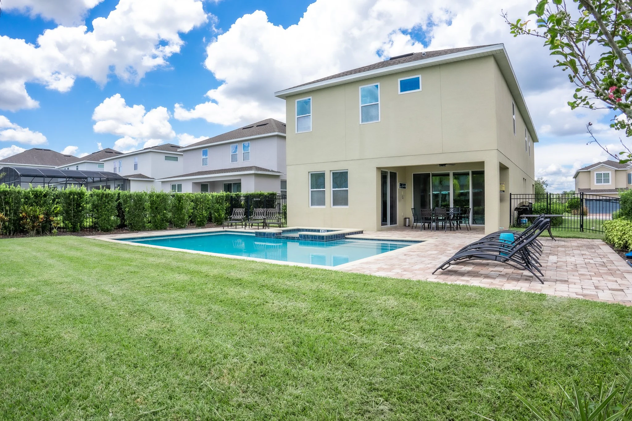 Backyard with swimming pool, patio furniture, lounge chairs, green lawn, and two-story house under a partly cloudy sky.
