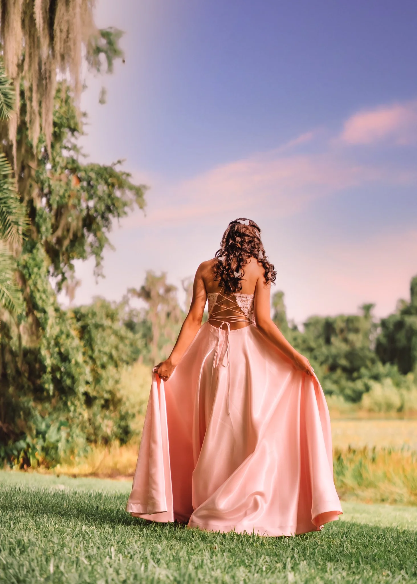 A woman in a pink satin gown standing outdoors in a grassy area, with her back to the camera, holding the sides of her dress. She has long, curly brown hair with a floral headpiece, and the background shows green trees and a blue sky with pink clouds