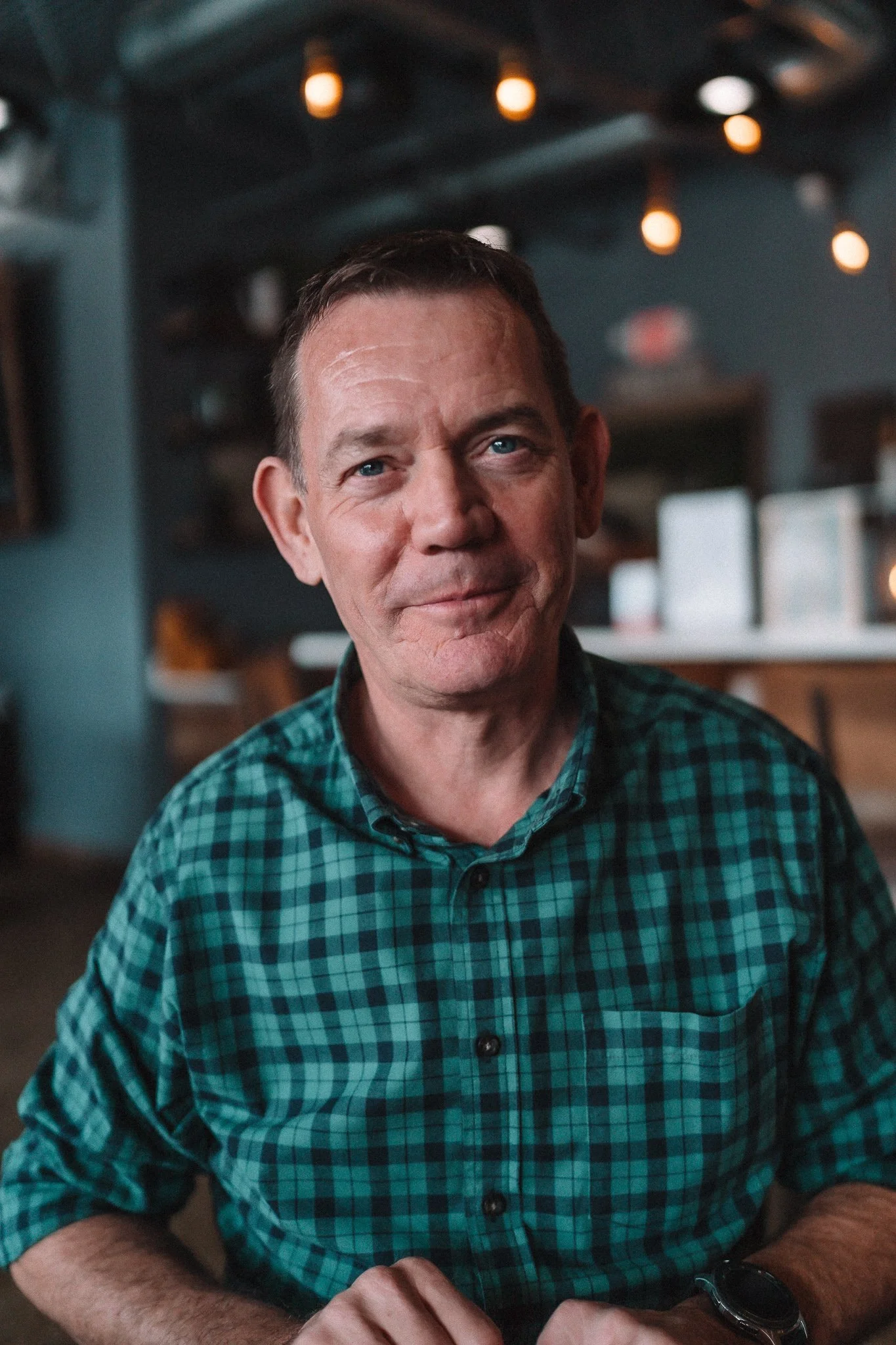 A middle-aged man with short brown hair, wearing a green and black checkered shirt, sitting in a dimly lit café or restaurant with warm hanging lights in the background.