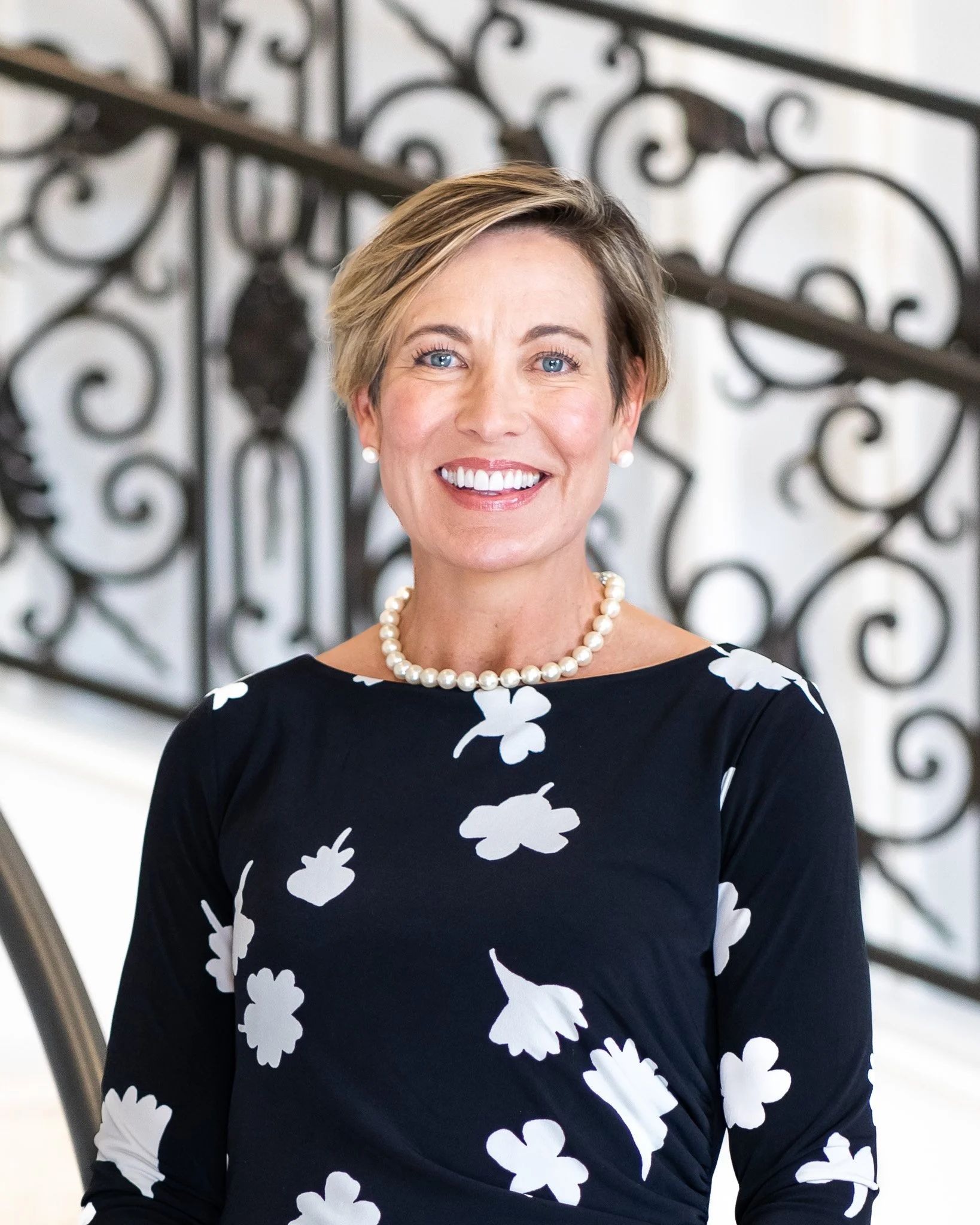 A woman with short blonde hair and blue eyes, smiling, wearing pearl earrings, a pearl necklace, and a black dress with white floral patterns. She is standing indoors near a decorative wrought iron staircase.