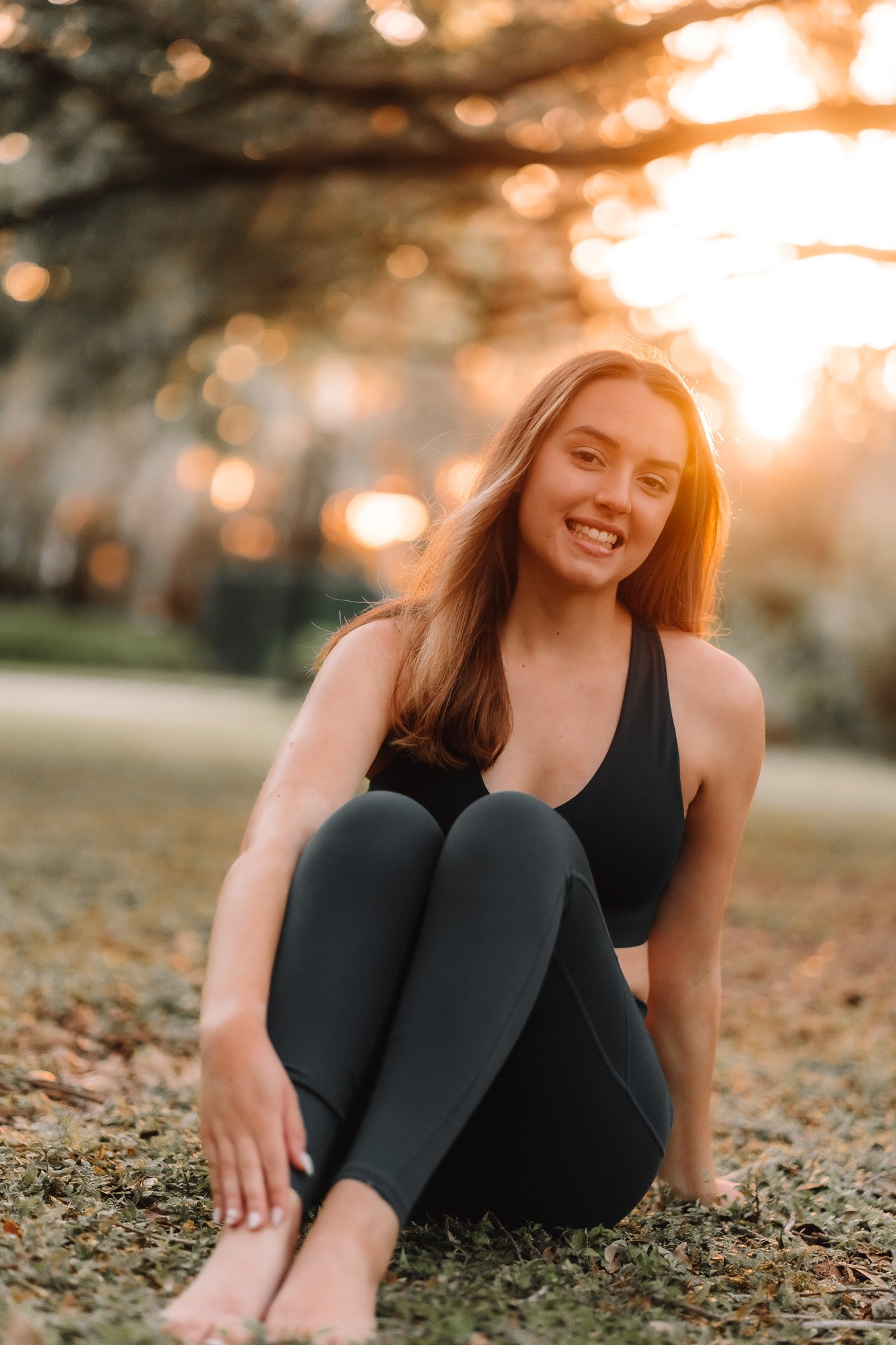 A young woman with long hair sitting outdoors on grass during sunset, smiling and wearing black athletic wear.