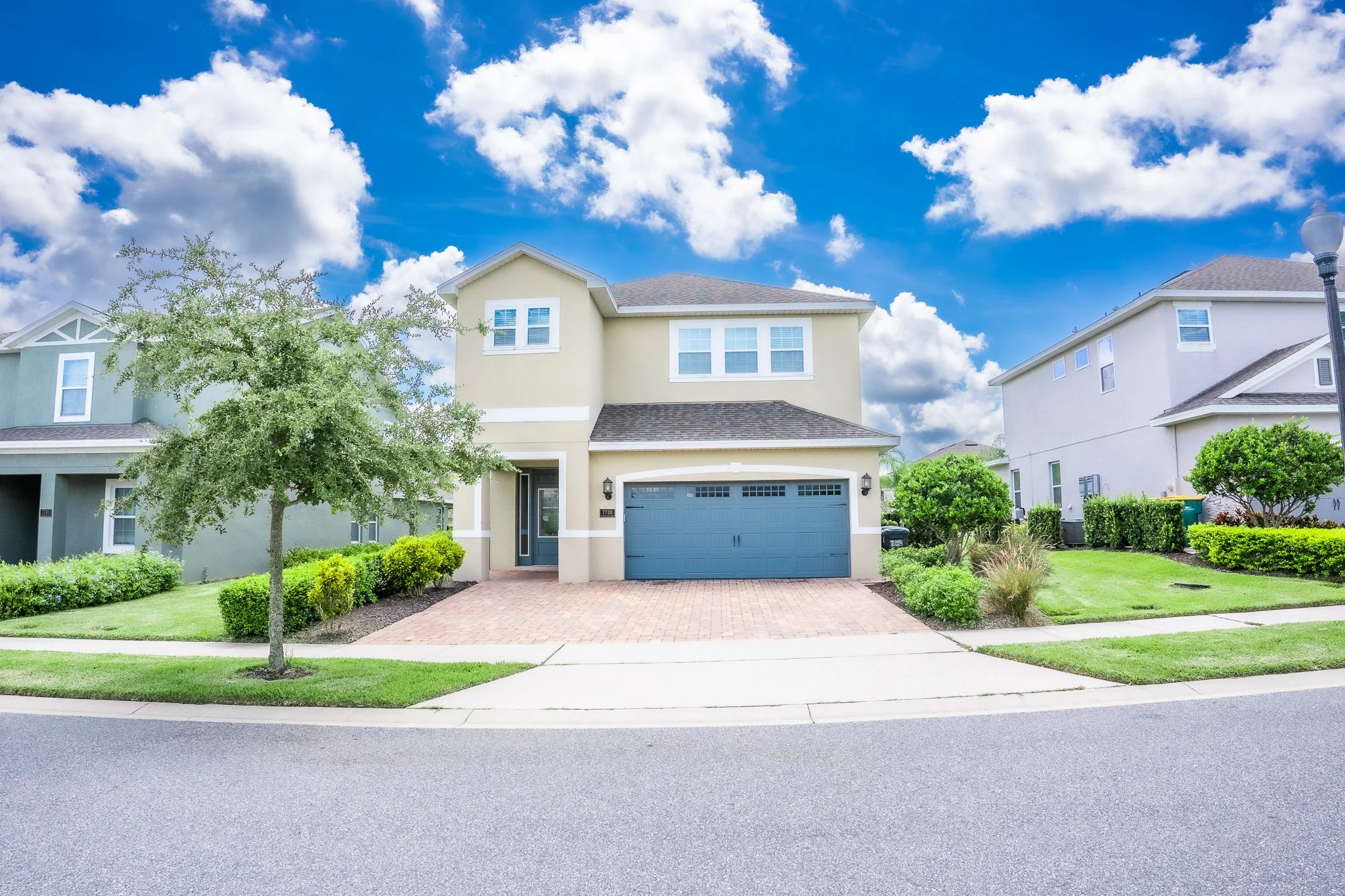 A two-story house with a blue garage door, beige exterior, and a small front porch. Well-maintained lawn with bushes and a tree, under a partly cloudy blue sky.