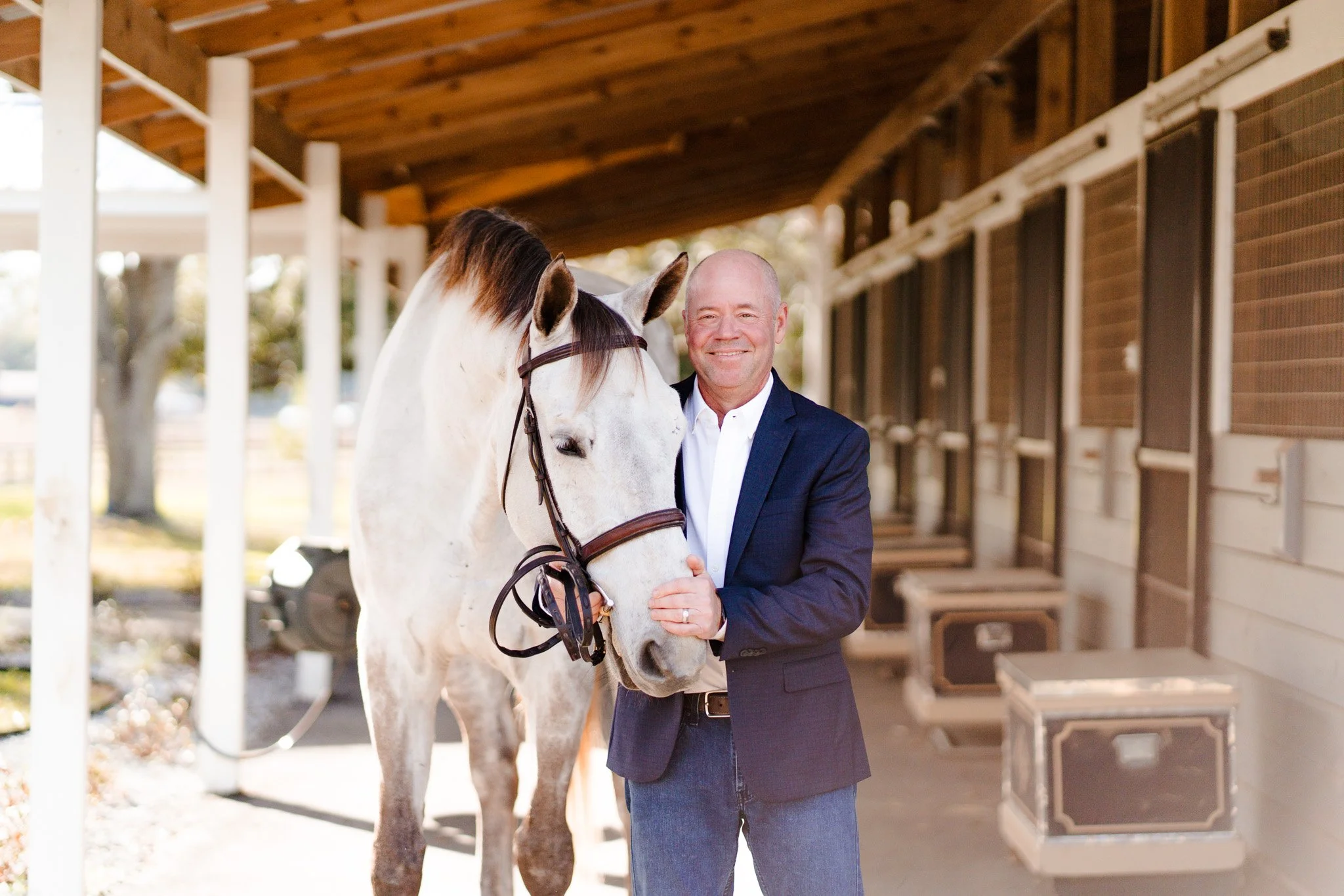 A smiling man in a suit standing next to a white horse in a stable area.