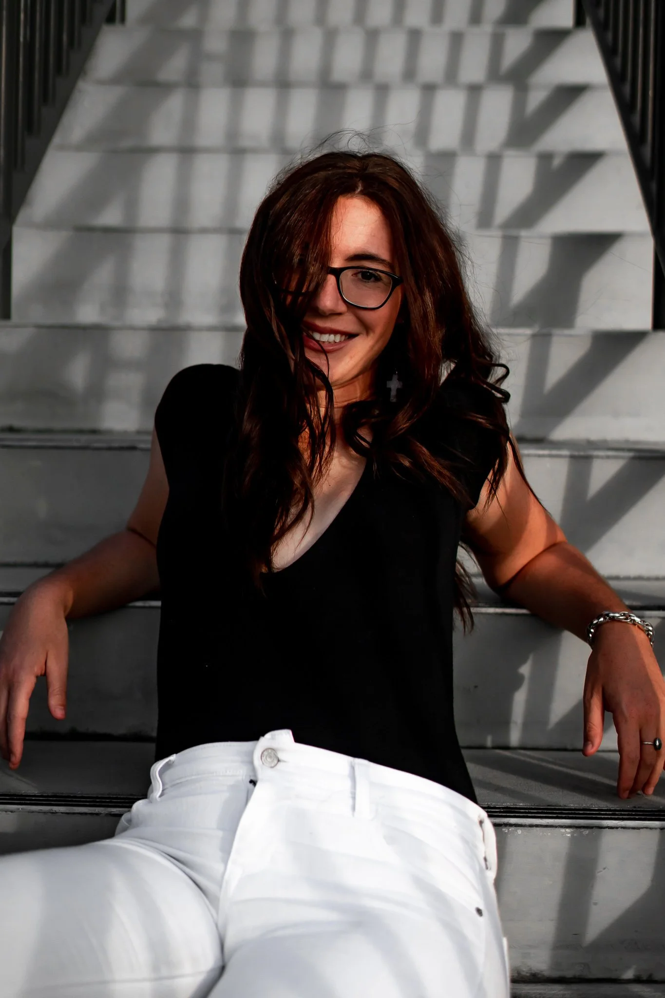 A woman with long dark hair, glasses, a black top, and white pants sitting on outdoor stairs, smiling at the camera.
