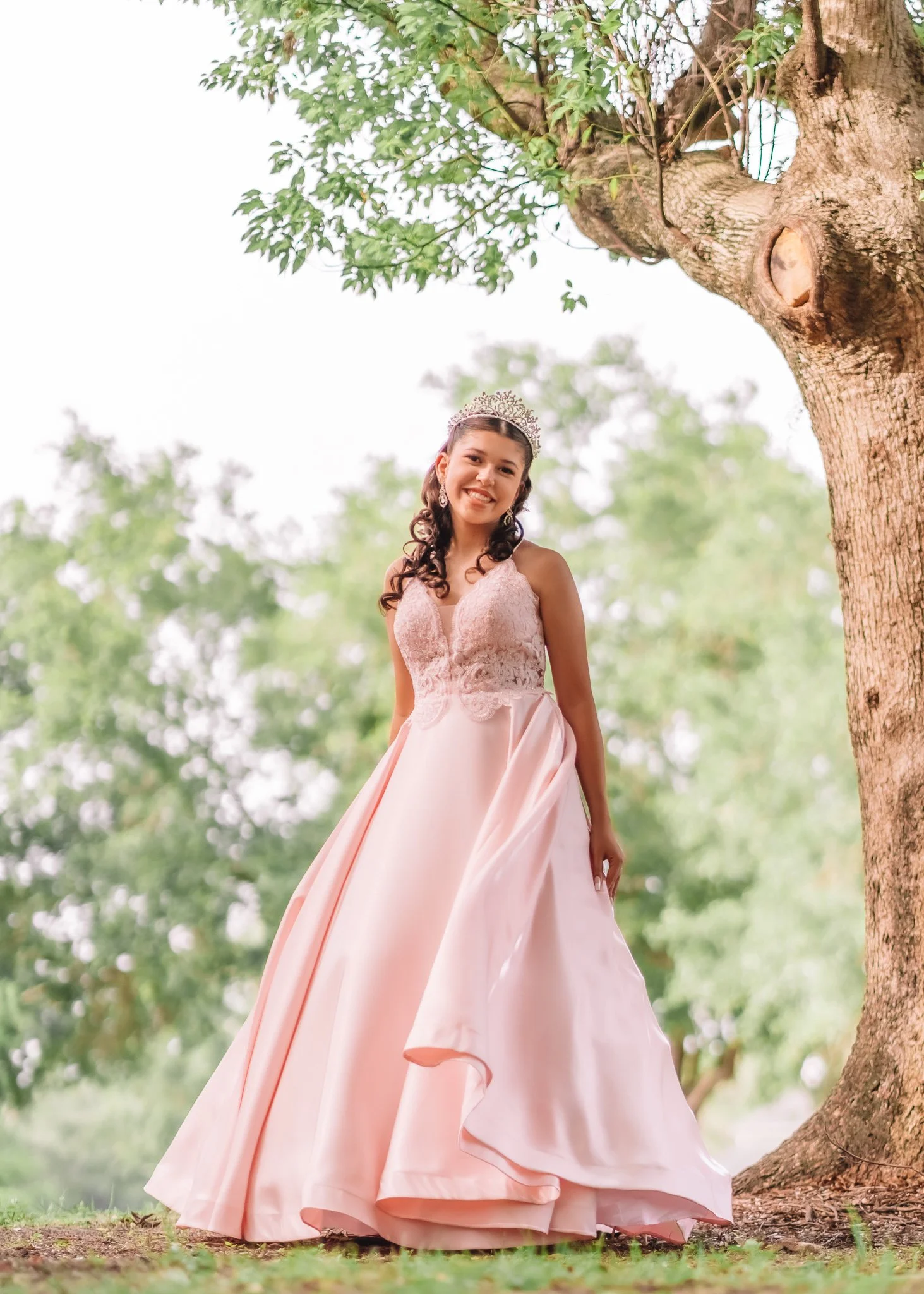 A young girl in a pink gown and crown standing next to a large tree with green leaves in an outdoor setting.