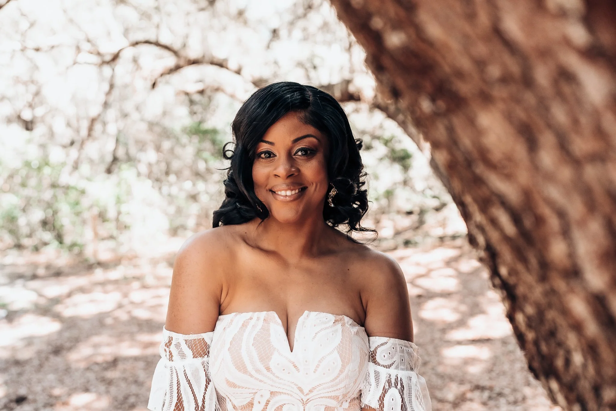 A woman with black hair and earrings smiling outdoors, wearing a white off-shoulder lace dress, surrounded by trees.