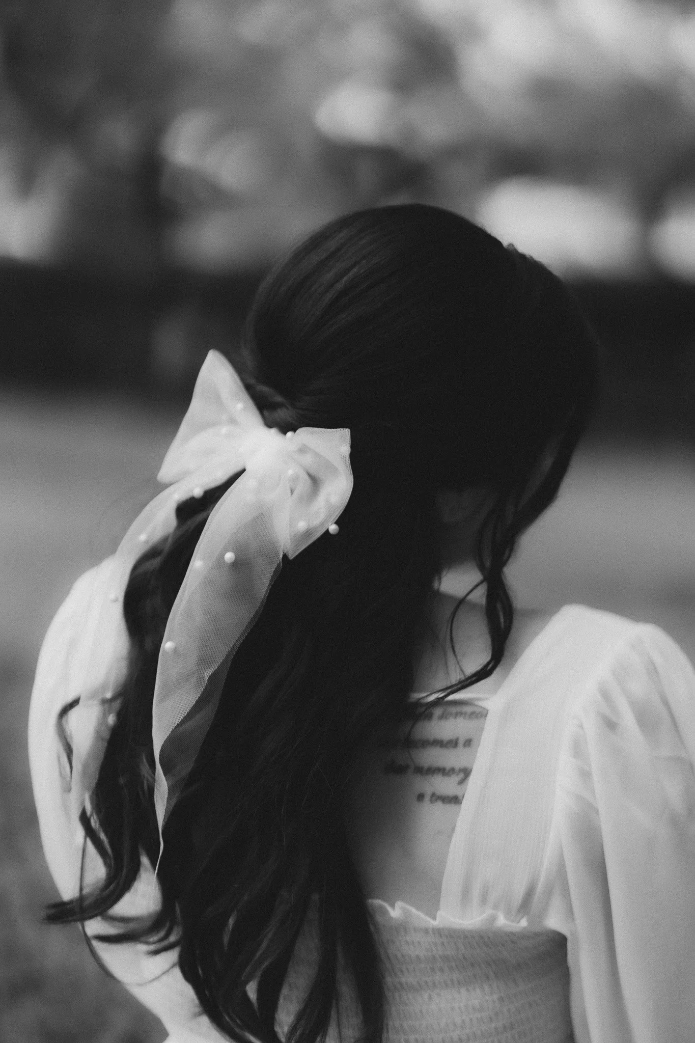 A close-up in black and white of a woman with long, wavy hair wearing a light-colored dress with lace patterns and a bow accessory in her hair, with a blurred background.