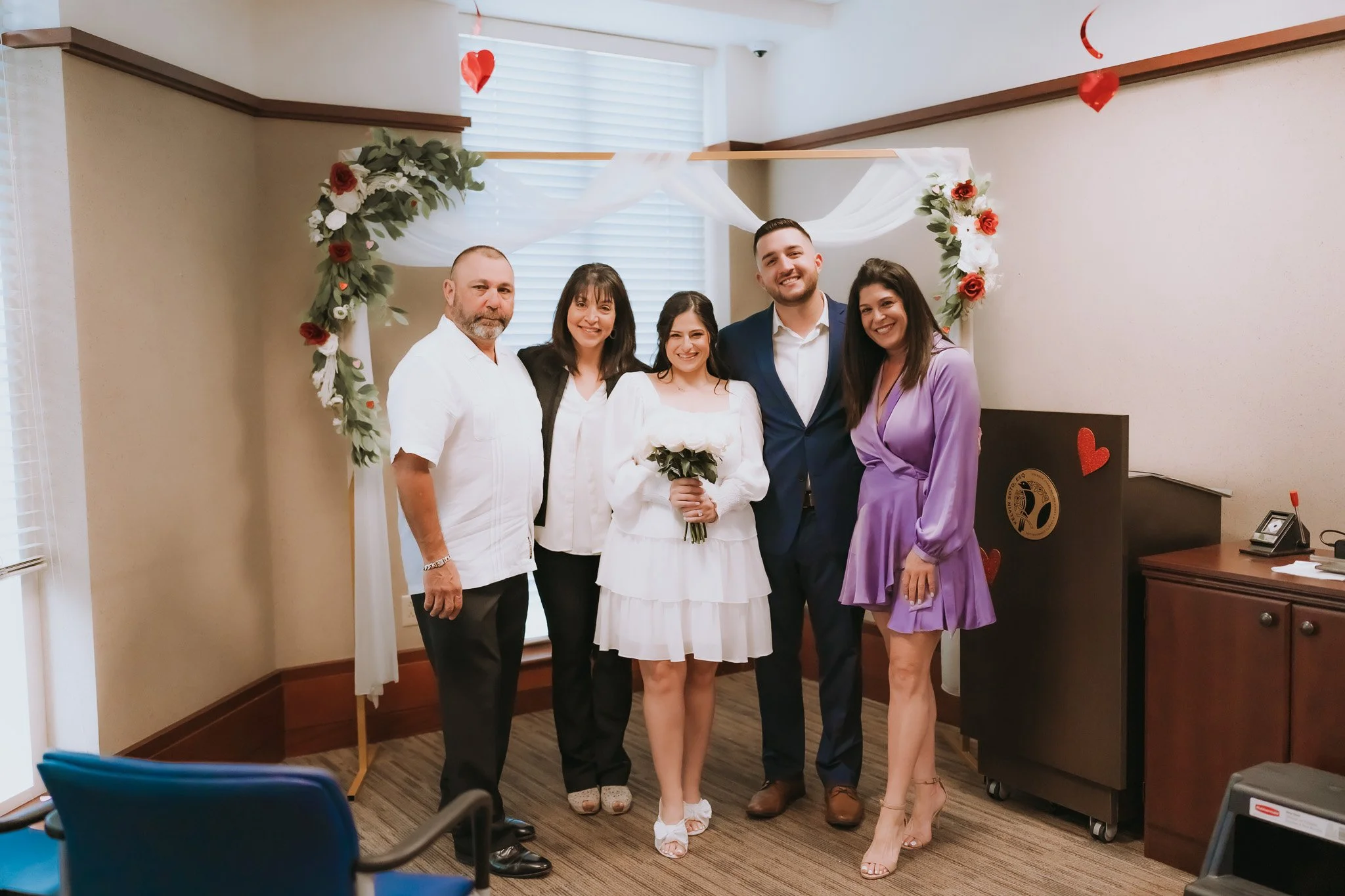 A group of five people standing together indoors in front of a decorated backdrop with flowers and tulle. The group includes three women and two men, all smiling and dressed for a formal occasion, possibly a wedding or celebration. One woman in the c