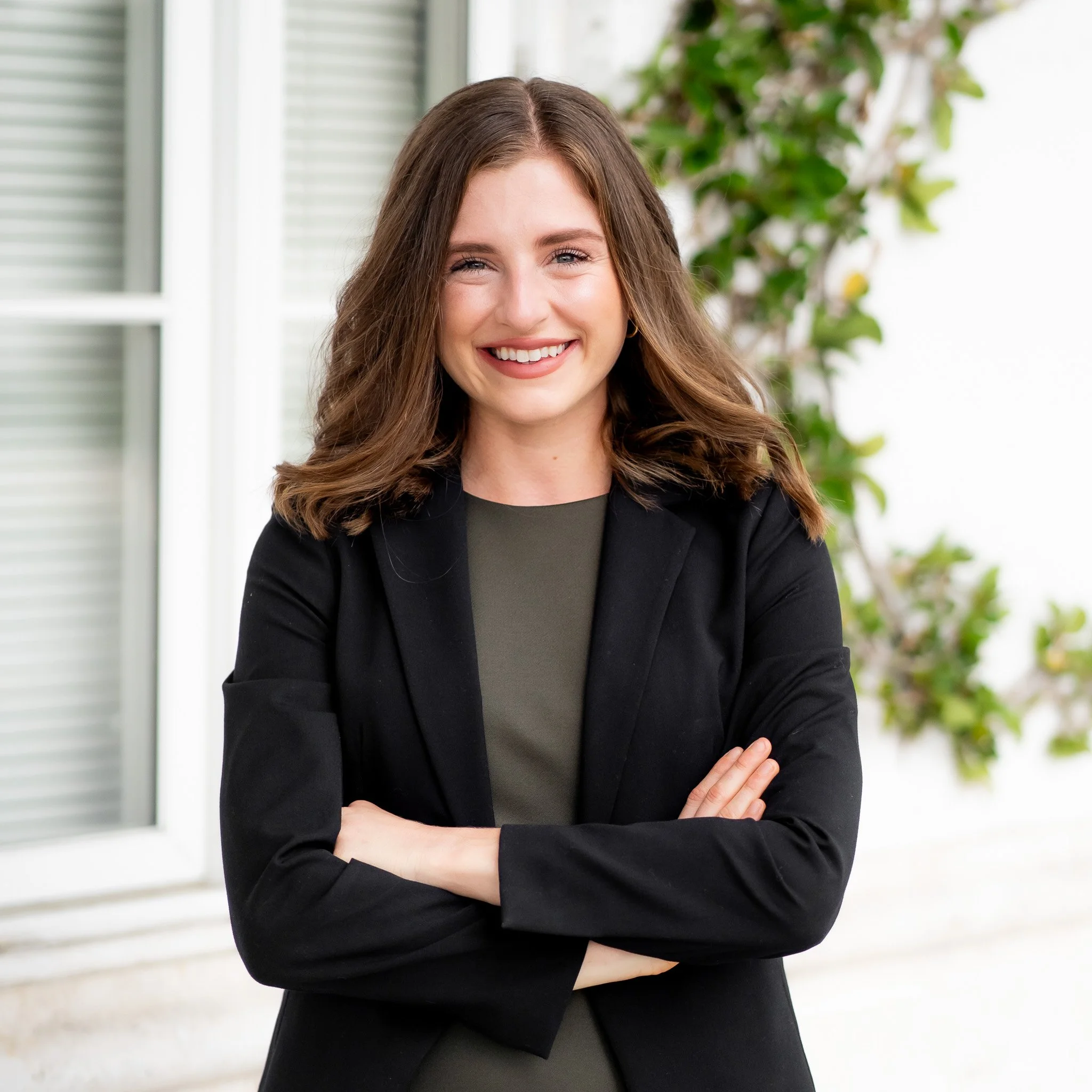 A woman with shoulder-length brown hair, smiling, wearing a dark blazer over a green top, standing outdoors with a background of a white wall, a window, and green plants.