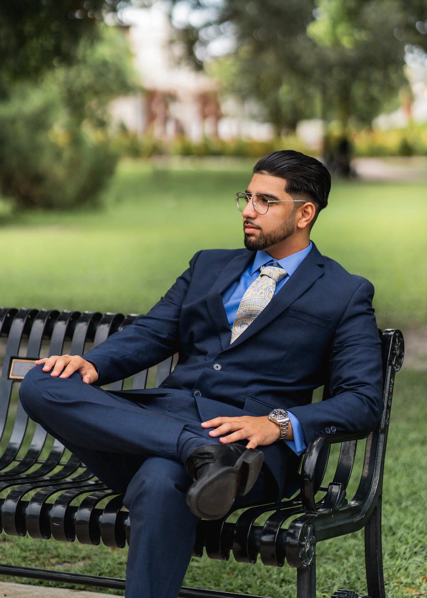 A man in a blue suit and tie sits on a park bench with one leg crossed over the other, wearing glasses and a wristwatch, outdoors on a sunny day.