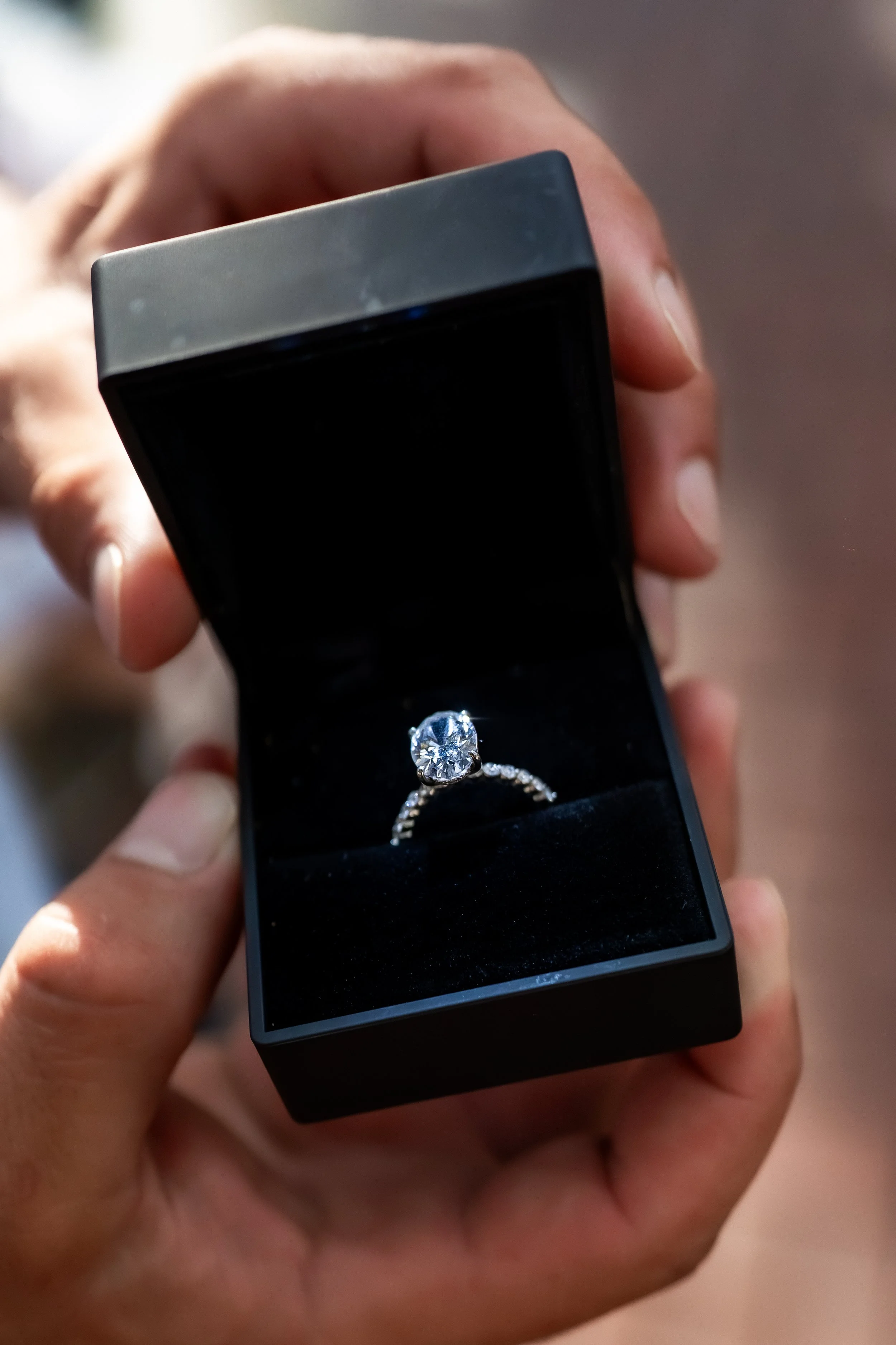 A person's hand holding an open black jewelry box with a diamond engagement ring inside.